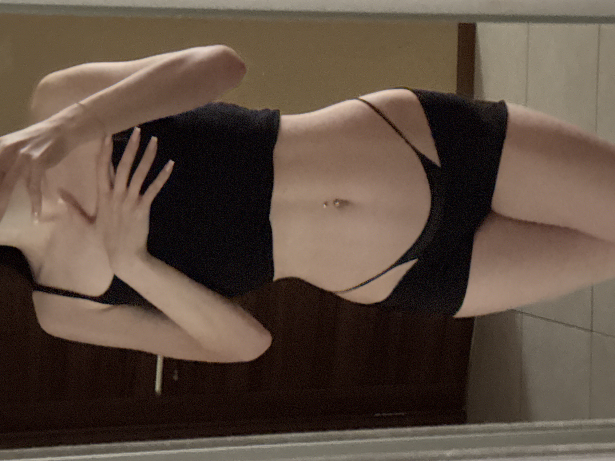 A person is standing in front of a mirror, reflecting their torso and upper legs. They are wearing a black crop top and black shorts with cutouts. Their hands are placed on their chest. The background appears to be an indoor setting with a dark wooden door to the left and a wall with a light brown color to the right. The flooring is tiled.