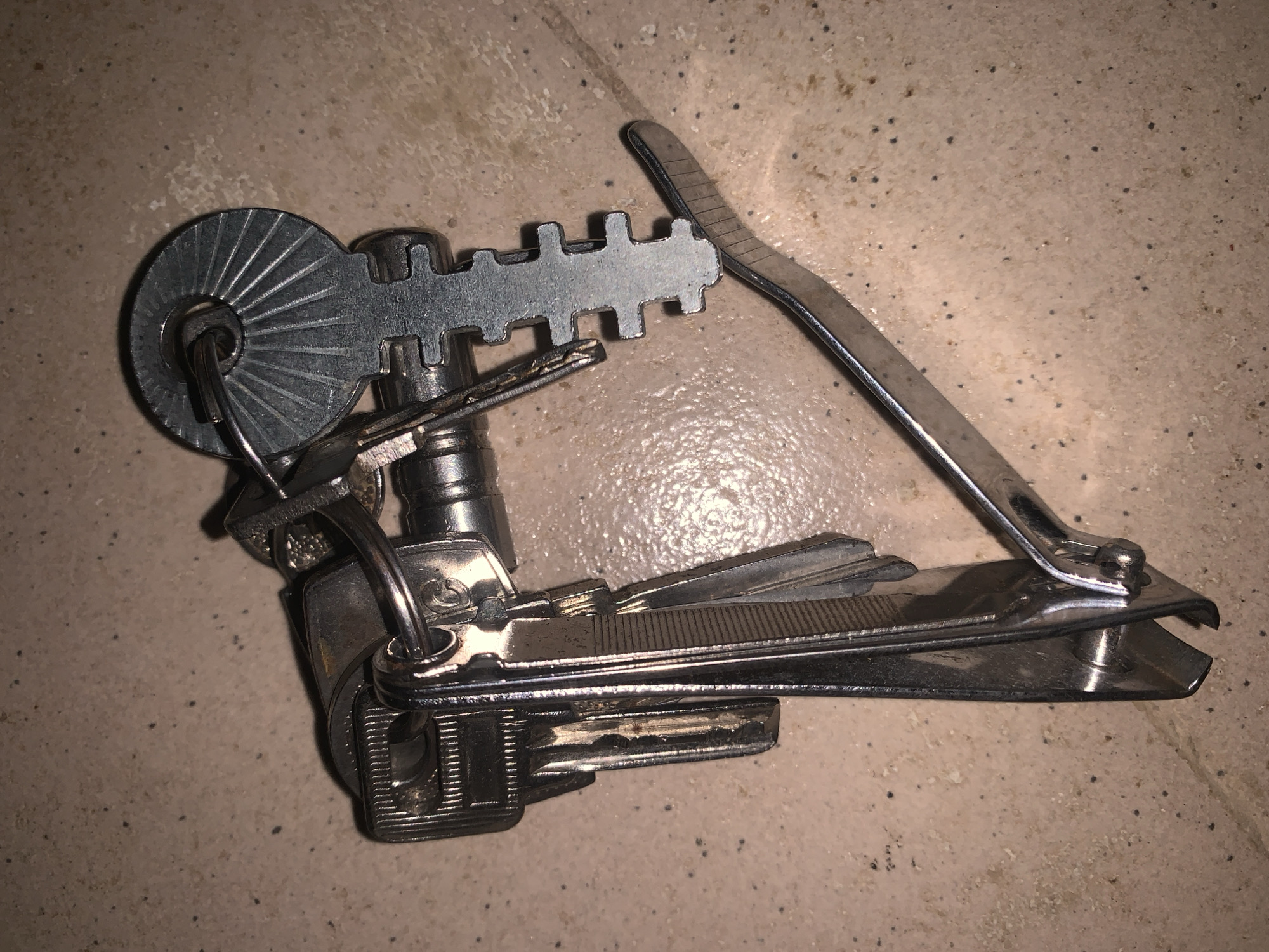 An overhead shot captures a cluster of silver keys and a nail clipper on a mottled beige surface. The keys are attached to a keyring, and one key has a distinct fan-shaped head with radiating lines. Another key has a notched, almost bone-like shape. Multiple other keys are partially visible. The nail clipper, also silver, is open, with its cutting blades positioned beneath the keys, and the lever is extended. The background is an indoor surface, possibly a countertop or floor, with dark specks scattered across it. The lighting is dim, creating shadows.