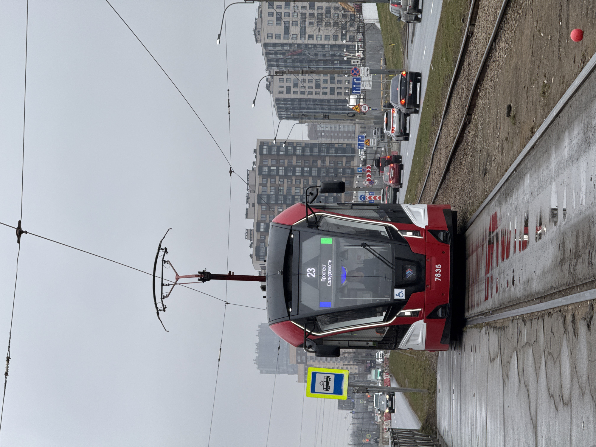 The image shows a modern red and black tram traveling along a tram line. The tram is labeled with the number 23 and the words "Проспект Солидарности", which translates to "Solidarity Avenue," indicating a location in Saint Petersburg, Russia. The tram's identification number "7835" is displayed on its front. The tram is equipped with a pantograph for drawing power from overhead wires. In the background, there are residential buildings and street lights. The weather appears to be overcast or rainy, as the street surface is wet and reflective. There are cars and other vehicles visible on the road alongside the tram tracks. A yellow sign with a tram icon is posted on the left side of the image, indicating a tram stop or route.