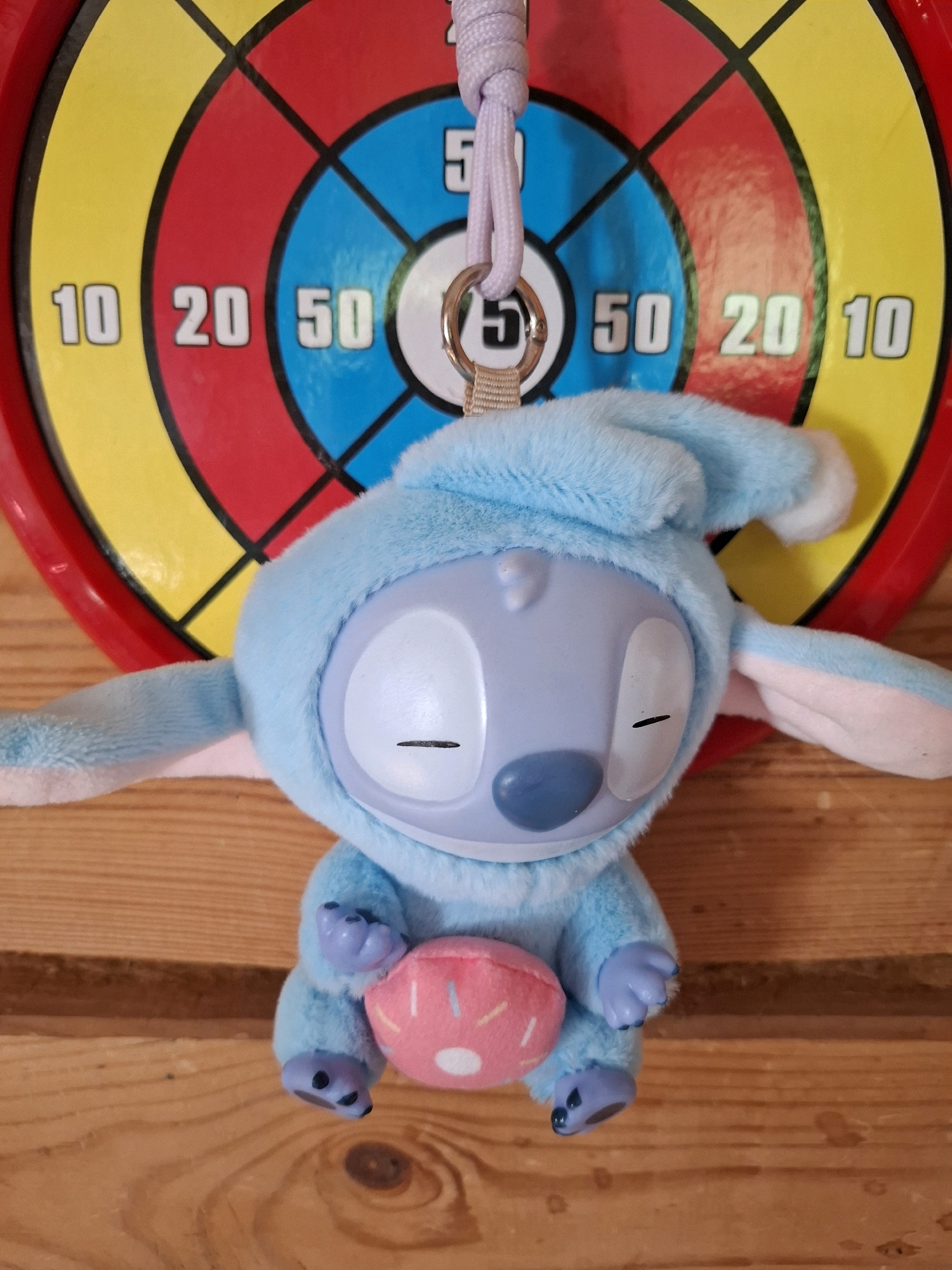 A plush toy of the character Stitch, dressed in a blue pajama outfit with a hood, is hanging from a purple string and a metal ring against a dartboard. The dartboard has concentric circles of red, yellow, and blue, with black lines dividing it into sections. Numbers are visible on the dartboard: "10", "20", and "50". The toy is light blue with large white eyes that have black horizontal lines for pupils, a small blue nose, and pink ear linings. It is holding a pink object with white circular dots, resembling a donut. The toy is suspended against a wooden surface, likely a wall or table. There is no indication of the location in Tyumen, Russia.
