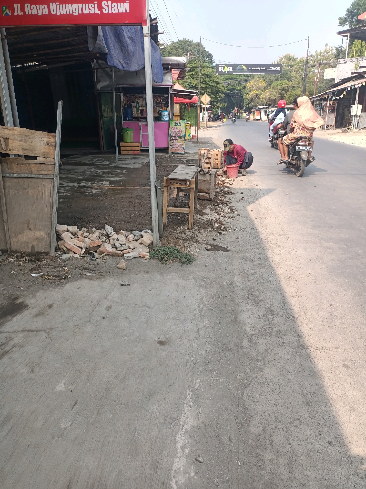 The image depicts a street scene in Ujungrusi, Slawi, Indonesia. The foreground shows a gray concrete sidewalk with scattered debris including rocks and broken bricks. To the left, a wooden structure with a weathered panel is partially visible. A person wearing a red shirt and a dark cap is squatting on the sidewalk, appearing to work with a red bucket and some wooden crates. Further down the sidewalk, a wooden bench is present. A motorbike is riding on the asphalt road, carrying two individuals; the rear passenger is wearing a light-colored headscarf and a patterned dress, and the rider is wearing a red helmet. Other motorbikes are visible further down the road, suggesting a moderately busy street. Shops and buildings line the street on both sides, with a prominent red sign at the top left that reads "Jl. Raya Ujungrusi, Slawi." A banner with the word "BLACK" is suspended over the road in the mid-ground. The environment appears to be outdoors, with trees and buildings under a clear sky.