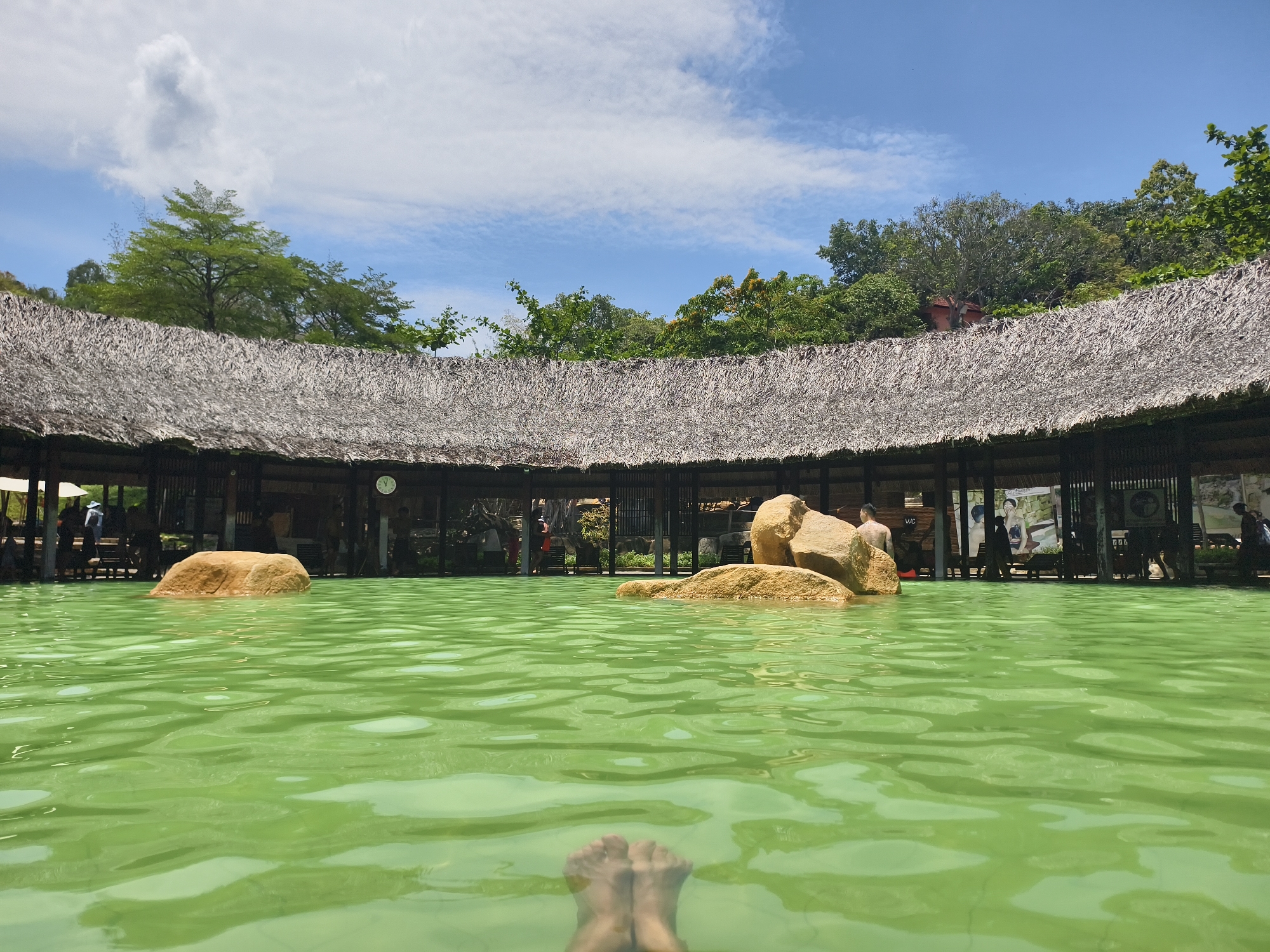 The image depicts a body of water, likely a pool or natural spring, with a distinctive green hue. Two large rocks protrude from the water's surface. In the foreground, a pair of feet are visible, suggesting the perspective of someone immersed in the water. The water's surface is rippled, indicating slight movement.
In the background, a semi-circular structure with a thatched roof is visible. The structure appears to be open on the sides, with columns or posts supporting the roof. People are present within the structure, some standing and others seated.
The sky is visible above the structure, with scattered clouds against a blue background. Trees and other vegetation are also visible in the background.
The location is Unknown, Unknown.
