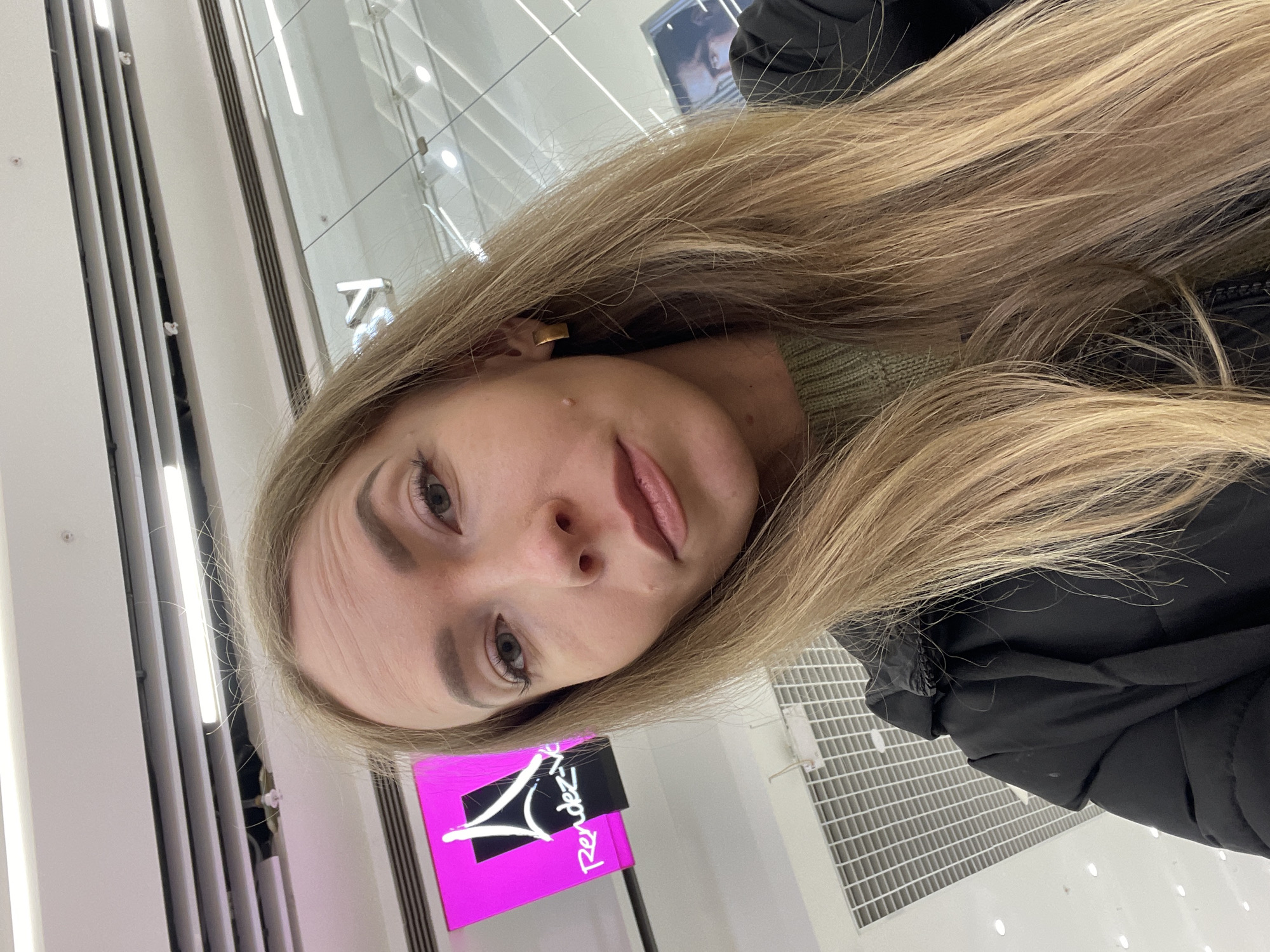A selfie of a fair-skinned woman with long blonde hair, wearing a green sweater and a black coat. She is indoors, likely in a shopping center in Perm, Russia, as evidenced by the presence of a "Rendez-vous" advertisement, a shoe store chain with locations in Perm. The background includes white walls with vertical panels and overhead lighting. A ventilation system is visible on the floor.