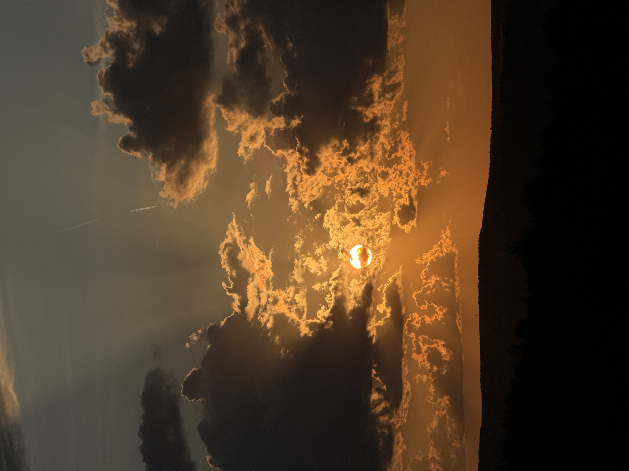 The image shows a dramatic sunset over a dark, silhouetted landscape. The sun, a bright orange orb, is partially obscured by dark, textured clouds that are illuminated from behind with fiery orange and yellow hues. Rays of light beam out from behind the clouds, creating a celestial display. In the distance, faint contrails from an airplane streak across the dusky sky. The foreground is a solid black silhouette of rolling hills and trees, creating a stark contrast with the vibrant sky above. The overall impression is one of awe and natural beauty, with the setting sun casting a dramatic glow.