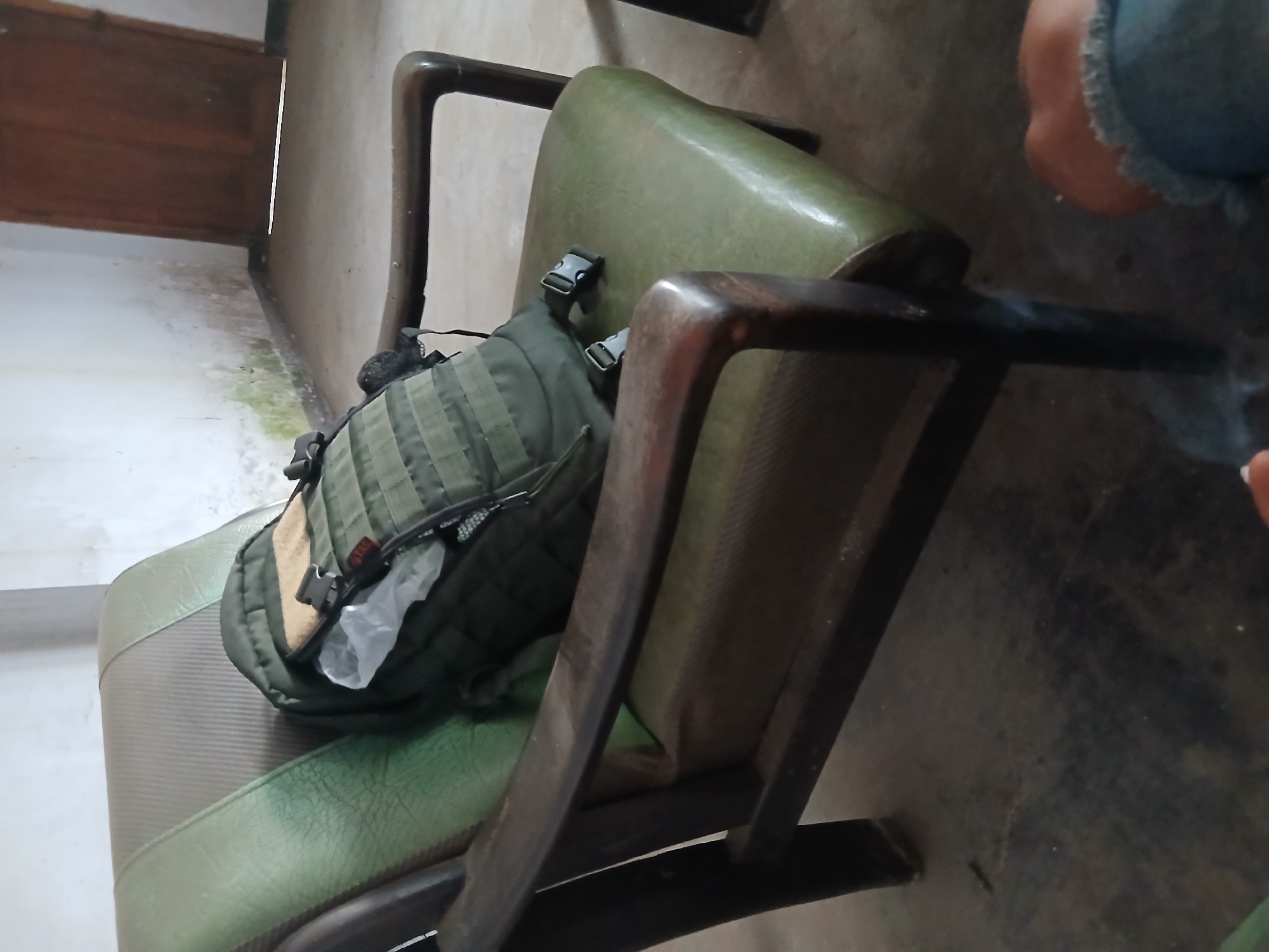 In a sparsely furnished room, a green leather armchair with a worn seat and ribbed backrest is occupied by a dark green tactical backpack. The backpack has multiple compartments and buckles, and a light-colored object, possibly a bag, is visible protruding from its opening. The armchair's wooden frame is visible, showing signs of age. The room's walls are a pale, textured color, with some areas exhibiting mildew stains near the baseboard. To the right, a dark wooden door stands ajar. The floor is concrete and appears dusty. In the foreground, the lower part of a person wearing blue jeans is visible, with their hand holding what appears to be a cigarette. The scene suggests a waiting area or a temporary stop, potentially in a public or semi-public space in Tenggarang, Indonesia, given the details of the room's construction and wear.