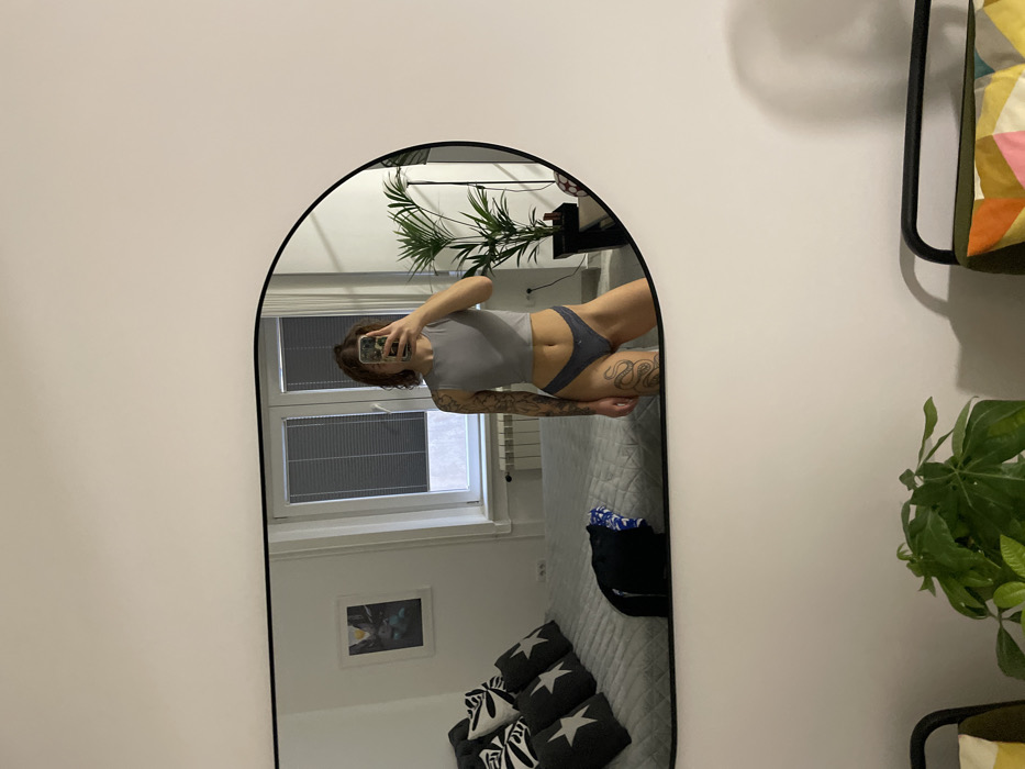 The image displays an indoor scene, likely a bedroom or studio apartment, captured as a reflection in a large, black-framed, rounded rectangular mirror mounted on a white wall. A female individual stands in the mirror's reflection, positioned centrally, holding a smartphone that obscures her face, indicating self-photography. She wears a grey sleeveless crop top and grey thong-style underwear. Her right arm is extensively tattooed, and a prominent tattoo, resembling a snake, is visible on her left thigh.
The reflected background reveals a large window with closed grey horizontal blinds, beneath which a white radiator is installed. To the left, a bed with a grey quilted spread and a dark headboard is visible, arranged with four pillows featuring black and white star patterns. A dark piece of clothing, with a visible blue and white pattern on its interior, rests on the bed. A framed artwork hangs on the wall above the bed. To the right of the individual, a tall, potted plant with dark green leaves stands on the floor.
In the unreflected foreground, a white wall is visible. A smaller potted plant with green leaves is positioned at the bottom left, and part of a black-framed chair or stool with an olive green and multi-colored geometric cushion is visible at the bottom right. The exact location within Belgrade, Serbia, cannot be determined from the visual evidence provided.