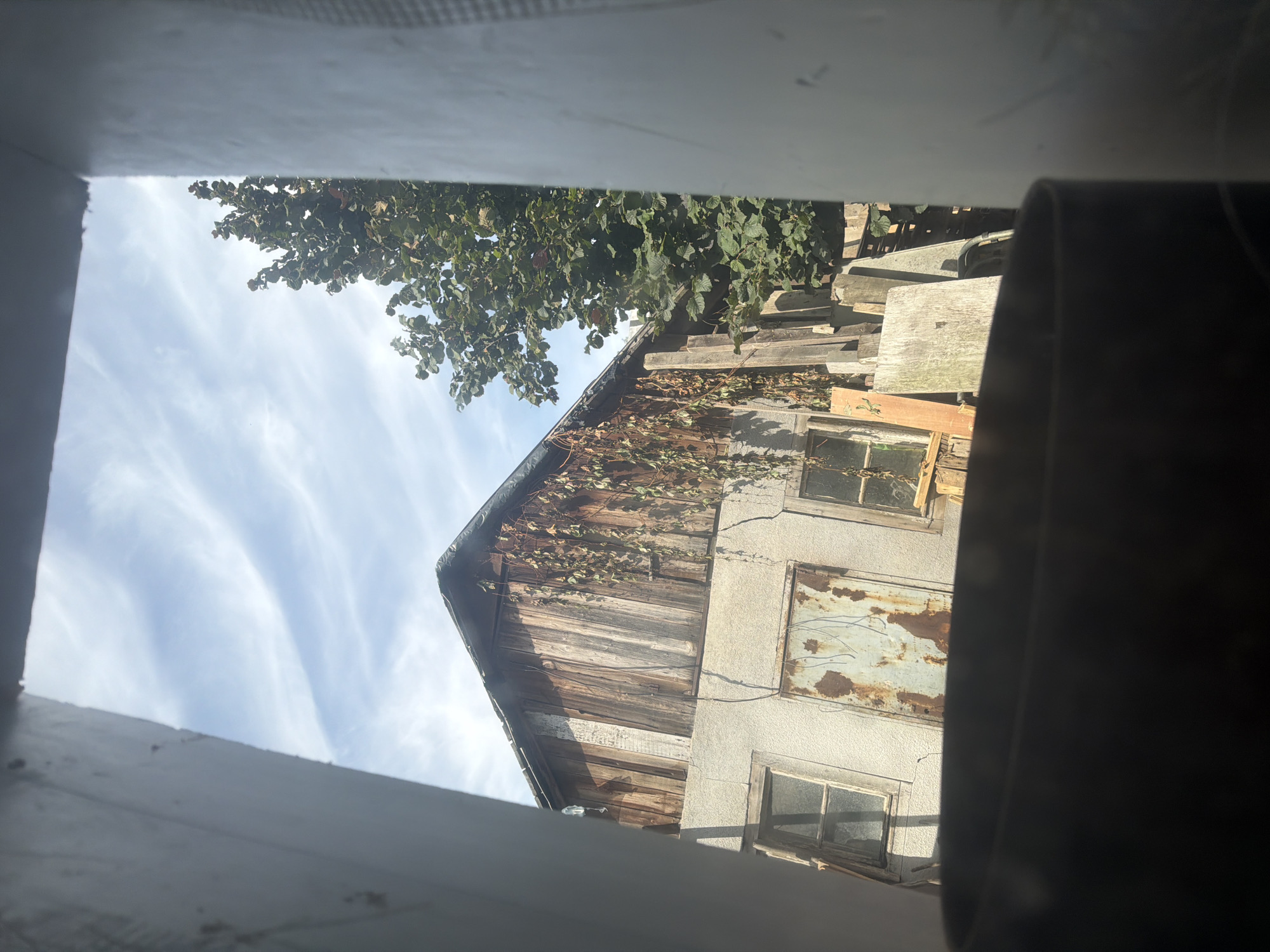 The image depicts a view through a dark, blurred foreground object, likely a container or pipe, looking out towards an old, weathered building. The building has a wooden upper section with a sloped roof covered in what appears to be dark tarp. The wooden planks are aged and some are covered in hanging vines. Below the wooden section, the building has a stucco-like lower level with a light gray, textured finish.
There are two visible windows on the lower level. The left window is rectangular and appears to have a broken pane. The right window is also rectangular and is partially obscured by hanging vines. Between the windows, there is a large, rectangular panel that appears to be a rusted metal door or cover, with visible streaks of rust and some scratched markings.
To the right of the building, a large tree with dense green foliage is visible, partially obscuring the sky. The sky above is blue with wispy white clouds. In front of the building, there are various pieces of lumber and wooden planks stacked or leaning against each other. The overall scene suggests an abandoned or neglected property.
The image does not contain any people or specific identifiable landmarks to pinpoint a location within Szczecin, Poland. The description is based solely on the visual elements present in the photograph.