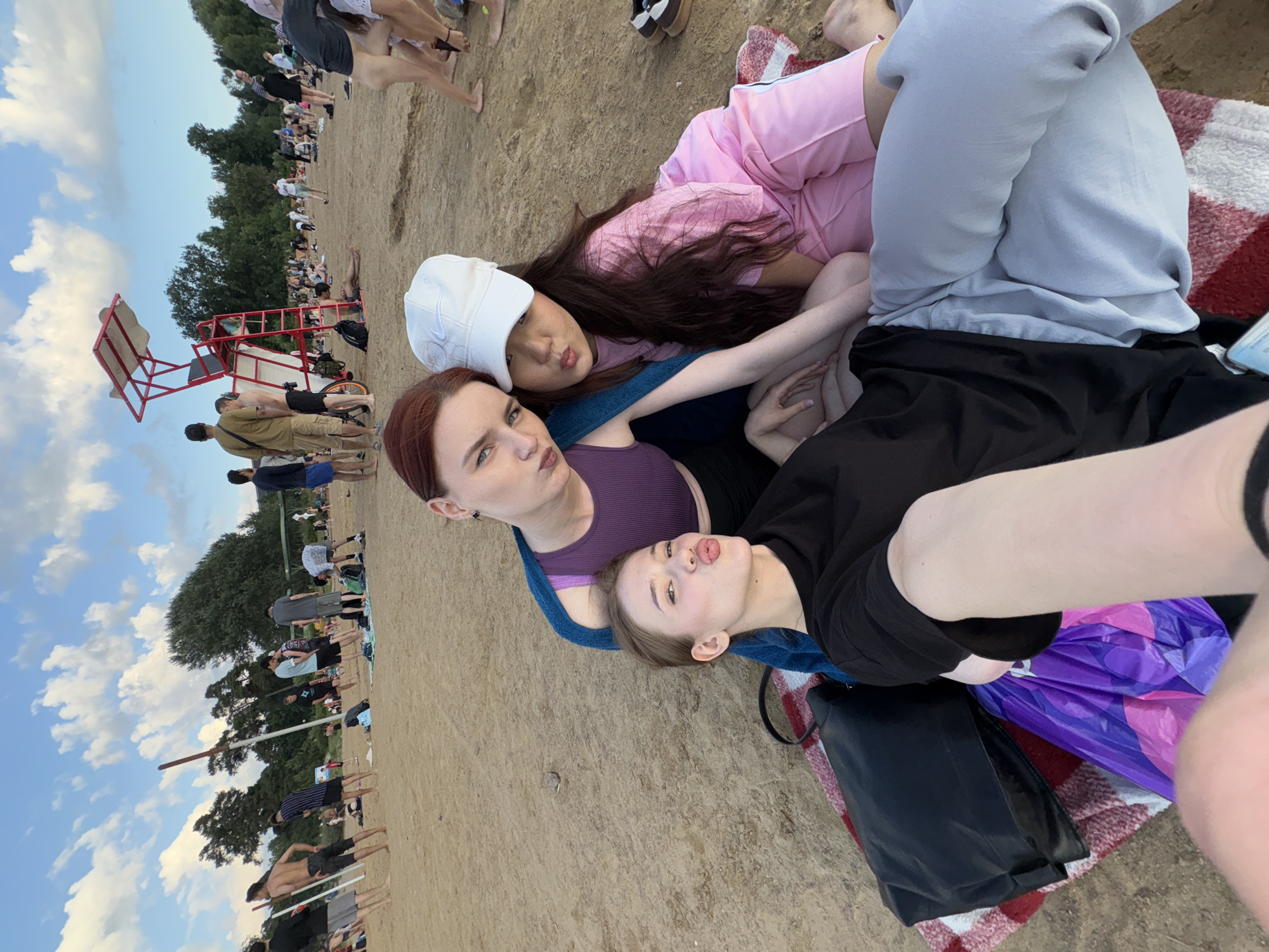 Three young women are lying on a picnic blanket on a sandy beach. The woman on the left has red hair and is wearing a purple top. The woman in the middle is wearing a white baseball cap and a pink outfit. The woman on the right is wearing grey sweatpants and a black shirt. All three appear to be posing for a photo, with two of them puckering their lips.
In the background, there are other people on the beach, some standing and some sitting. There is a lifeguard stand with a red frame and a white top. Trees line the edge of the beach, and the sky is blue with white clouds. The scene appears to be a casual gathering on a warm day, likely in the afternoon. The overall mood seems relaxed and playful. No text is visible in the image.
