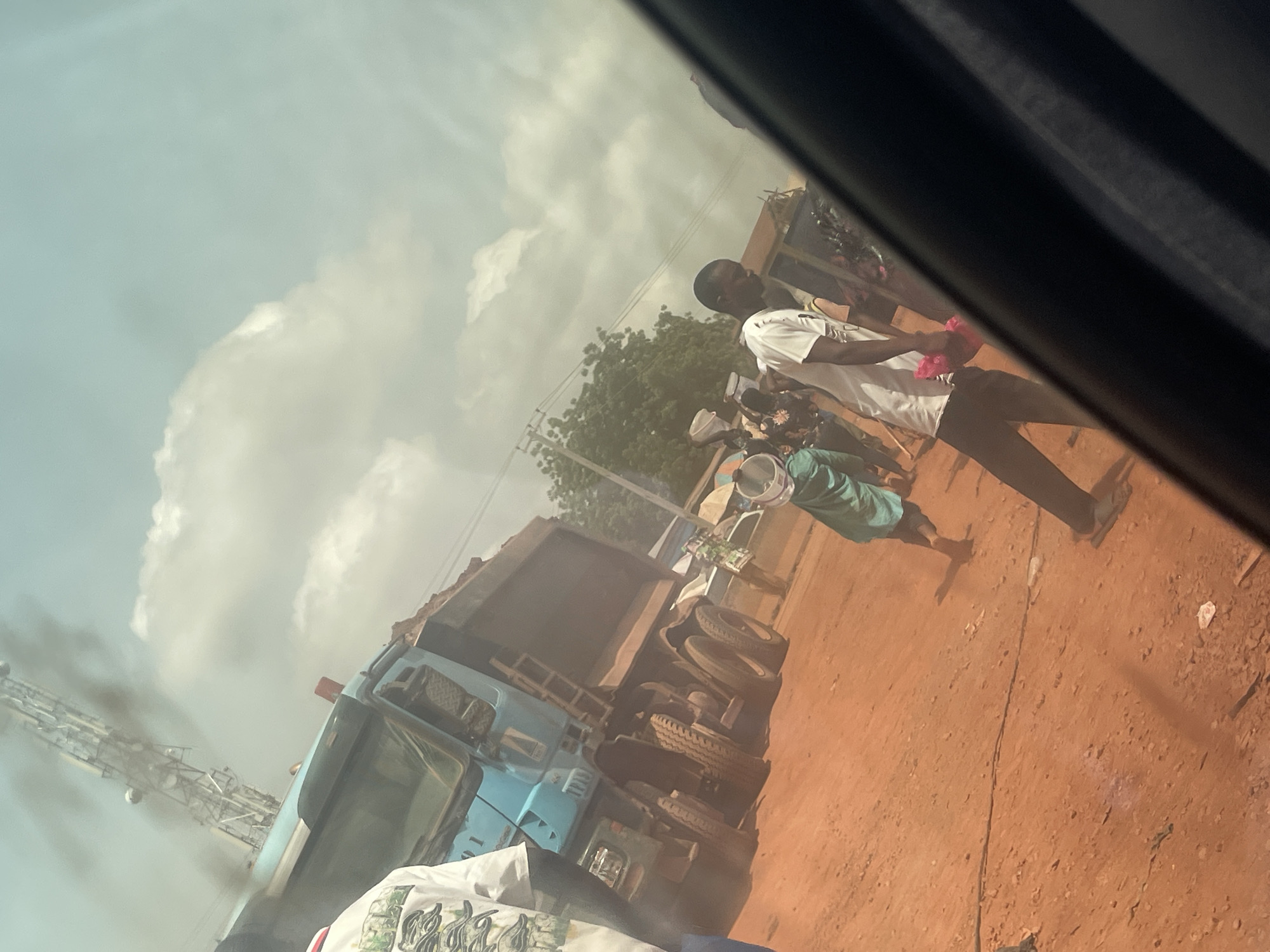 This image captures an outdoor street scene in Ado, Nigeria, from the perspective of someone inside a vehicle, looking out through a window. The photograph is tilted, showing a diagonal horizon.
In the immediate foreground, part of the dark interior of the vehicle is visible along the top and right edges, with some glare or reflection present. On the bottom left, a crumpled piece of white fabric, possibly a bag or clothing, can be seen.
The ground is a distinctive reddish-brown, consistent with laterite soil, and appears to be a dusty, unpaved road or shoulder. Several people are present; one prominent figure is a man in a white short-sleeved shirt and dark trousers, standing and looking towards the left side of the frame. Another person, partially visible in a teal or green top, is holding a white bucket. Other indistinct figures are in the background.
A large light blue or teal dump truck occupies a significant portion of the mid-ground, appearing to be stationary or moving slowly. Its bed seems to contain a load, possibly sand or gravel. In the distance, on the far left, a tall communication tower or antenna structure is visible against the sky.
The sky is bright with scattered white clouds, suggesting it is daytime, likely mid-morning or afternoon. There is a noticeable haze or dust in the air, contributing to a somewhat soft and diffused lighting, especially around the ground and truck. The overall impression is a bustling roadside environment in a semi-urban or rural setting. No legible text is clearly visible on any objects or clothing.