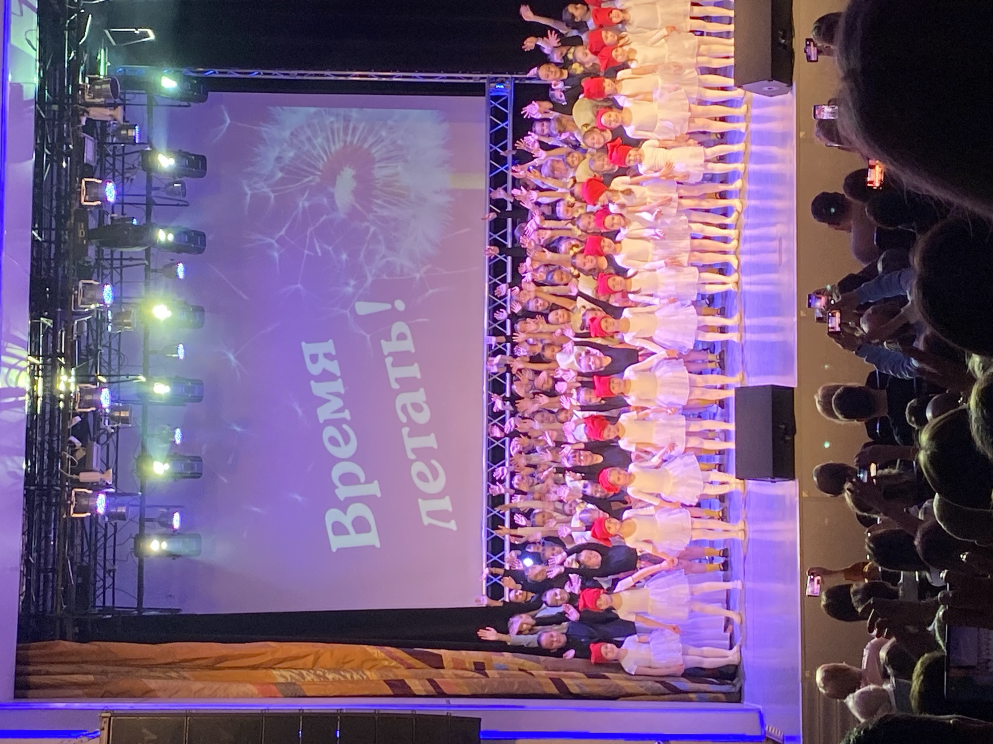 This image captures an indoor performance taking place on a stage, likely in an auditorium or theater in Chaikovsky, Russia. The scene is dominated by a large group of young girls, appearing to be children or pre-teens, performing on the stage. Most of the girls are dressed in white dresses or tutus, and many wear distinctive red caps. They are arranged in rows and appear to be engaged in a synchronized dance or theatrical routine, with many having their arms raised towards the audience.
Behind the performers, a large projection screen serves as a backdrop. The screen displays a graphic of a dandelion and Cyrillic text that reads: "Время летать!" which translates to "Time to fly!". To the left of the stage, several stage lights are visible, mounted on trussing, illuminating the performance area with various colors.
On the right side of the image, a section of the audience is visible, watching the performance. Many audience members are holding up smartphones, actively recording the event. The lighting in the venue suggests an evening or night-time event, with the stage brightly lit and the audience area appearing dimmer. The atmosphere appears to be one of engagement and excitement, typical of a children's performance or concert.