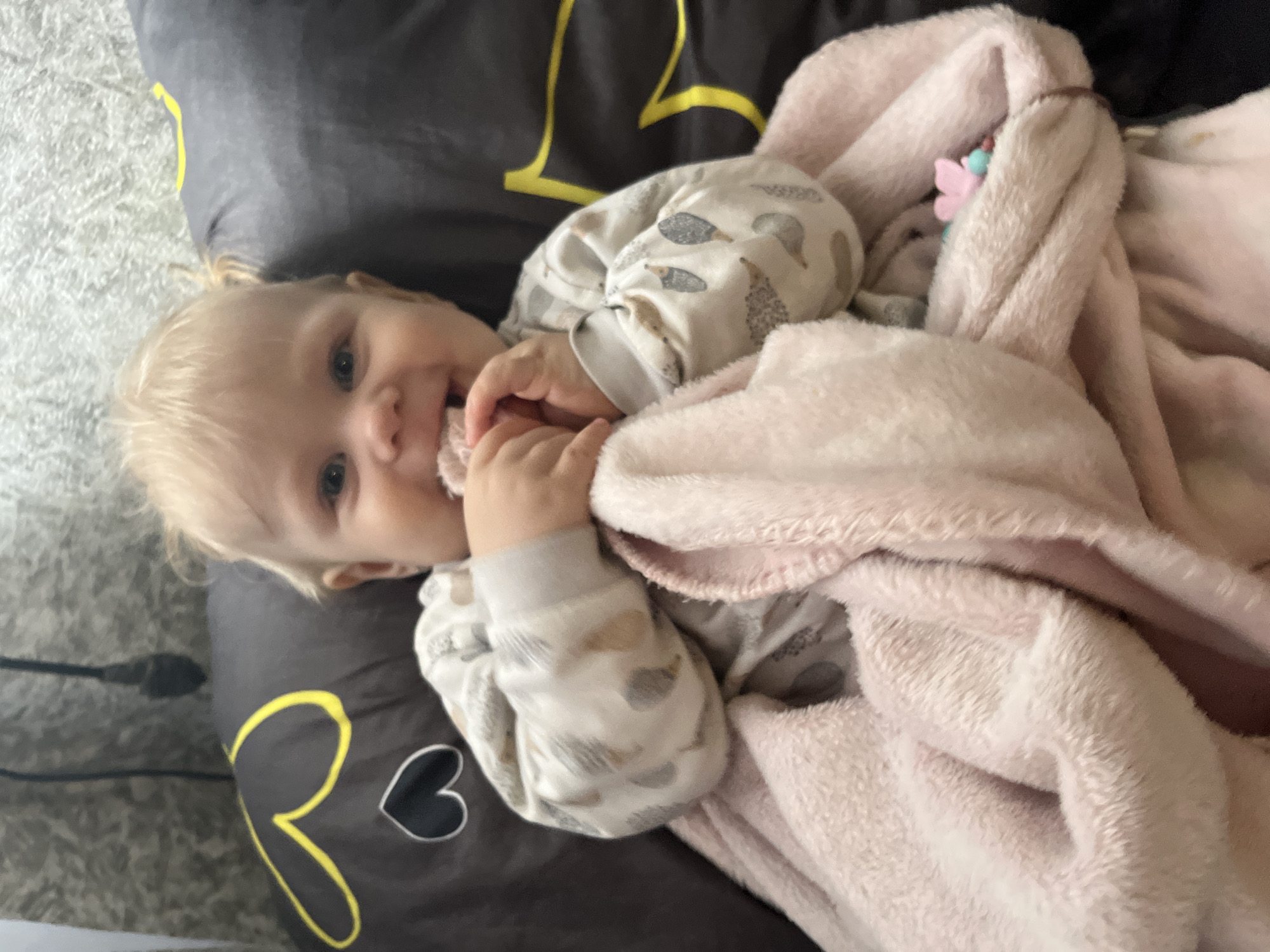 An indoor, eye-level shot captures a light-haired baby lying on a dark gray surface with a soft, pink blanket covering its lower body. The baby, with light blonde hair and blue eyes, looks directly at the camera, smiling with a joyful expression. Its hands are positioned near its mouth, possibly chewing on its fingers or the blanket. The baby wears a light-colored sleepsuit or pajamas patterned with small hedgehogs in shades of gray and brown.
The dark gray surface underneath the baby features a large yellow letter 'R' and a black heart outline. To the left of the baby, a light-colored, textured floor or rug is visible. A black cable extends from the bottom left corner across the textured surface. Tucked into the pink blanket near the baby's head is a small, multi-colored object, possibly a toy or pacifier attachment, featuring a pink butterfly shape. The scene suggests a relaxed, playful moment, likely during the daytime. The location context is Фоки, Russia.