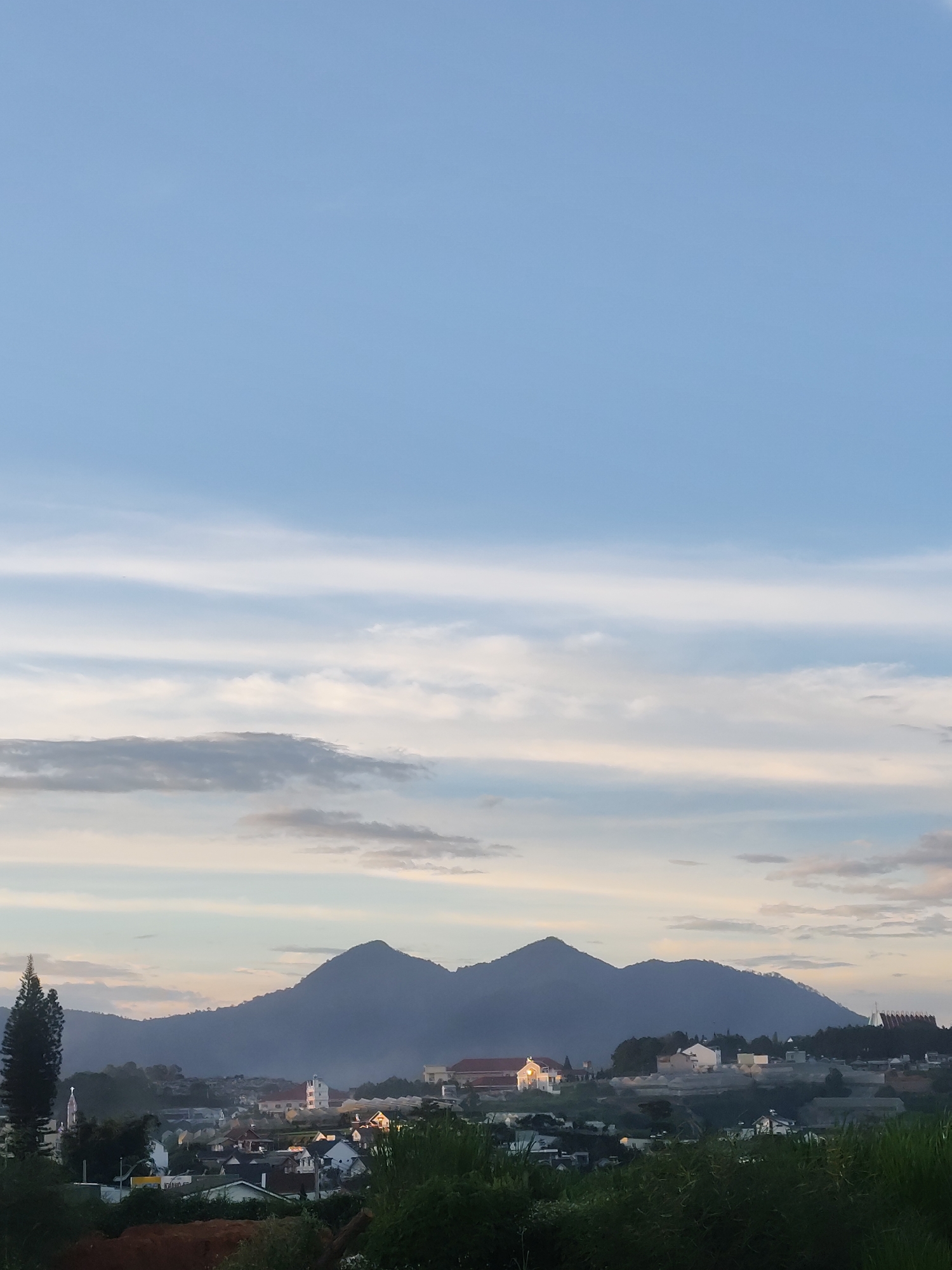 This image captures a serene landscape view of a residential area nestled in the hills of Da Lat, Vietnam, with a prominent mountain range in the background. The sky is a soft blue with scattered wispy white clouds, transitioning to softer hues near the horizon. In the foreground, lush green vegetation partially obscures the view of houses with varied architectural styles, some featuring white facades and red-tiled roofs. A tall, slender pine tree stands to the left, its dark green foliage contrasting with the lighter tones of the scene.
Further into the mid-ground, several structures are visible, including what appears to be a church with a white steeple and a larger building with a distinctive red-tiled roof. The mountainous backdrop is cloaked in a bluish haze, with two distinct peaks forming the central silhouette. The overall impression is one of tranquility and a picturesque semi-urban environment against a natural, undulating terrain.