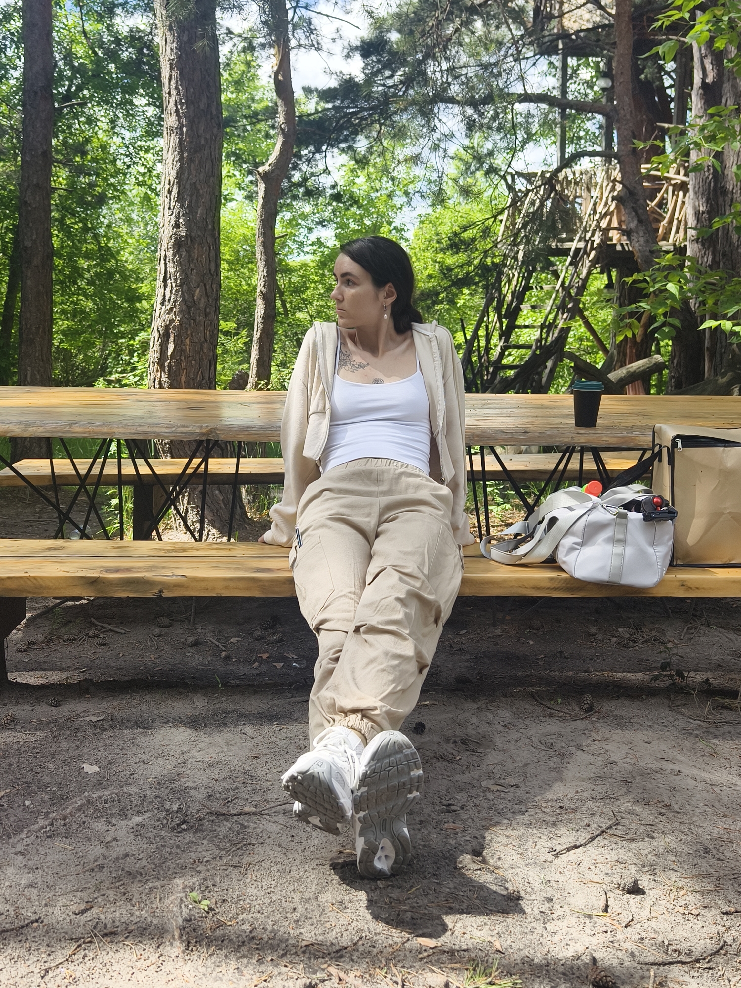 A young woman with dark hair is sitting on a wooden picnic table bench in a forest setting. She is wearing a white tank top, a cream-colored cardigan, and tan cargo pants. Her legs are crossed and her white sneakers are visible. To her right on the bench are a black disposable coffee cup, a white tote bag, and a brown paper bag. The bench is made of light-colored wood and has black metal supports. The ground is covered in dirt, pine needles, and scattered pine cones. In the background, there are tall trees with green foliage and a wooden structure resembling a treehouse. The scene appears to be a recreational area within a forest, likely located in the vicinity of Onochino, Russia, given the natural environment.