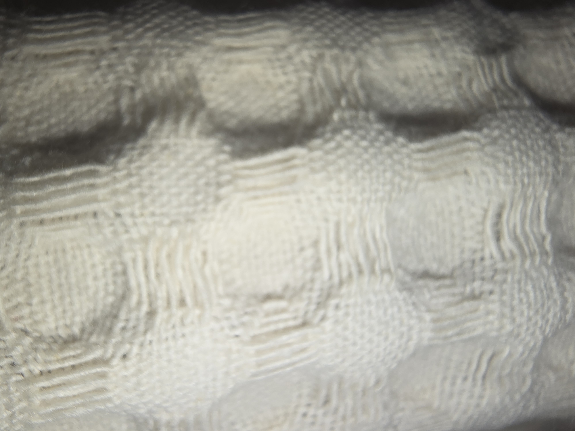The image is a close-up, blurry, and abstract shot of a textured, white fabric. The fabric appears to be knitted or woven, with a repeating pattern of interlocking loops or stitches that create a grid-like effect with raised areas. The lighting is dim, with some areas appearing darker and casting shadows, suggesting the fabric may be draped or folded. There are no people or discernible objects visible, and the background is dark and indistinct. The location of this image in Ulan-Ude, Russia, cannot be determined from the visual information provided.