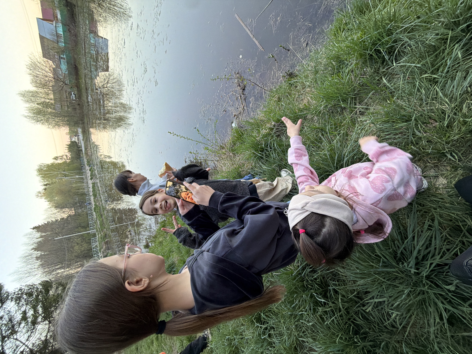 A group of children is lying on a grassy bank next to a body of water. The location is likely in Chaikovsky, Russia, based on visual cues of architecture and landscape.
Specifically, four children are visible. One girl with long brown hair and glasses faces the others. A girl in a black jacket holds a cell phone, seemingly taking a photo or video. Another child is eating. A girl in a pink hooded sweatshirt and a white headband is lying face down, with one arm raised. The children appear to be interacting and enjoying time together by the water.
The background includes a body of water, likely a river or lake, with vegetation along the bank. Distant buildings and trees are visible on the opposite shore. The light suggests it might be dusk.