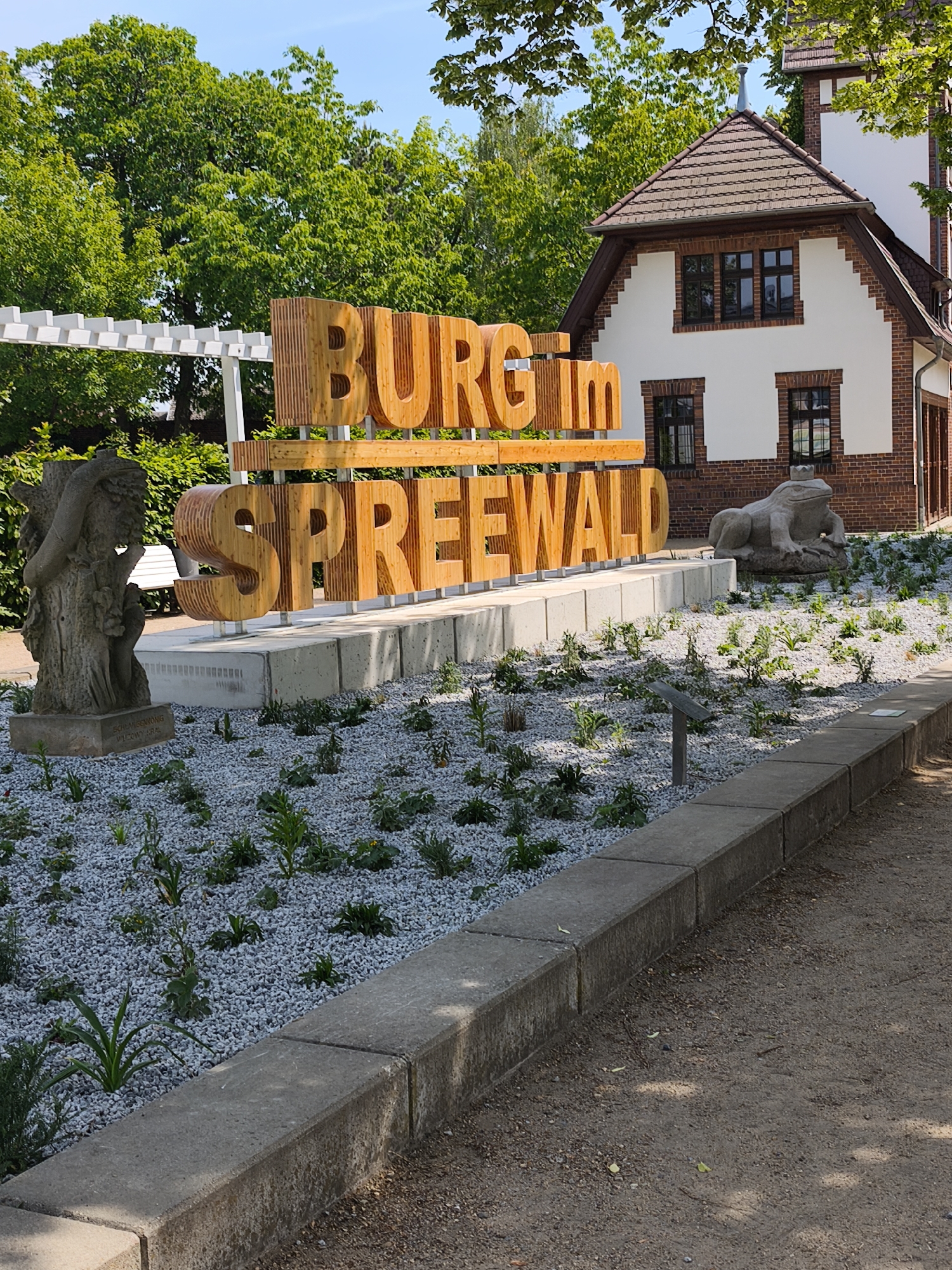 The image depicts a sign that reads "BURG Im SPREEWALD" in large wooden letters, located in an unknown city. The letters are installed on a concrete platform within a garden area covered with small white stones and scattered greenery. To the left of the sign, there is a stone statue, and to the right, a stone sculpture of a frog wearing a crown. In the background, there is a white building with dark brown window frames and a tiled roof, partially obscured by green trees. A pergola structure is visible to the left of the sign, behind the statue. The foreground consists of a gray concrete curb separating the garden area from a dirt path.