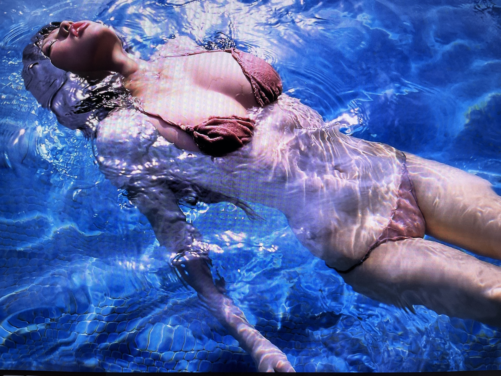 A woman is floating on her back in a swimming pool with blue water. Her eyes are closed, and her head is tilted back. She is wearing a pink bikini. The water is clear, and the bottom of the pool is visible. The scene appears serene and tranquil.