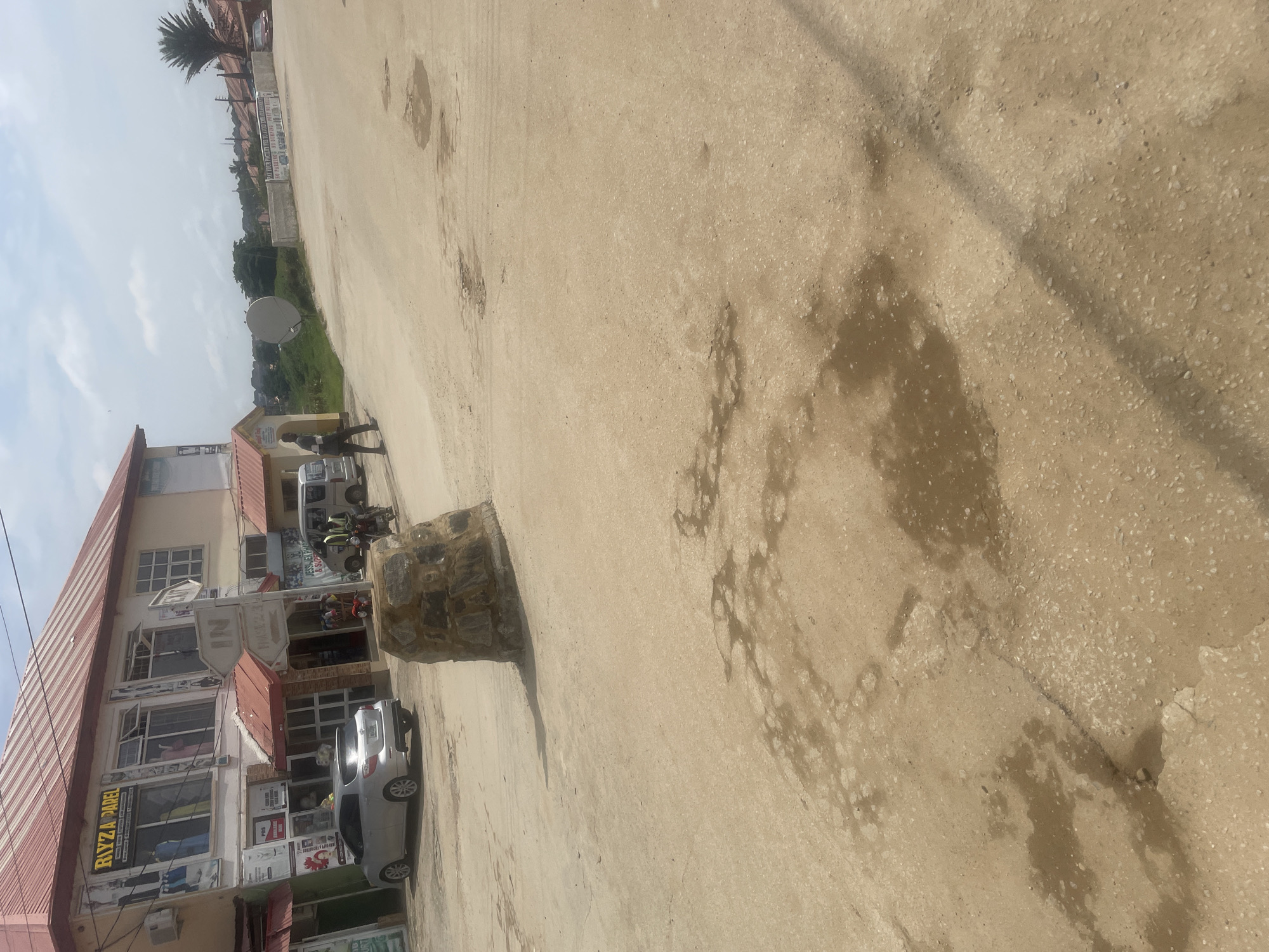 This is a vertical, wide-angle photograph taken in Abuja, Nigeria. The scene depicts a street or open area in front of a multi-story building. The ground is paved with a light-colored, textured material, with some areas showing darker patches that appear to be water stains or oil slicks.
In the foreground and midground, a large, irregularly shaped rock or stone structure is prominently placed on the ground. To the left of the rock, a silver car is parked. Further down the street, another silver vehicle, possibly an SUV or minivan, is also parked. There are several people visible in the midground, standing near the parked vehicles and the building. One individual appears to be wearing dark clothing and is walking. Another person is standing near a motorbike.
The building on the left has a reddish-brown roof and multiple windows. Several signs are visible on the building's facade, though the text is difficult to read clearly from this angle. One sign with yellow lettering reads "RIYZA PAREL." Another sign features the letters "NI" in a stylized font. A satellite dish is mounted on a pole in the background. The sky is partly cloudy. The overall impression is a daytime scene in a semi-urban or commercial area. The lighting suggests it is daytime, possibly late morning or early afternoon.