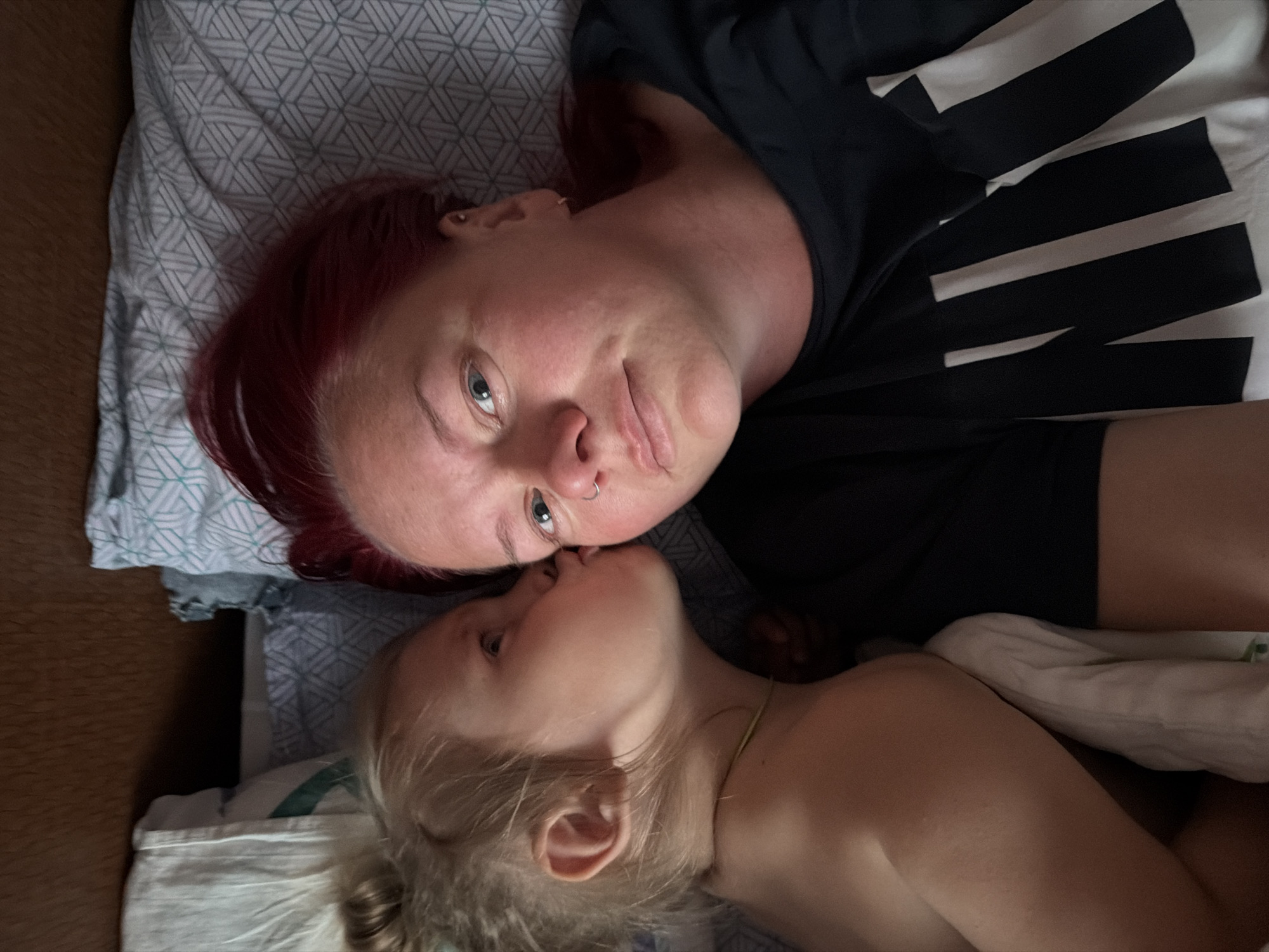 The media file shows a close-up, overhead view of two people, likely a mother and child, lying down together on a bed. The person in the foreground has blonde hair and is leaning in to kiss the person behind them. The person behind has red hair and is looking towards the camera with one eye visible. They are both wearing dark clothing, with the person in the back wearing a black t-shirt with large white stripes on it. The scene is indoors, with patterned bedding visible. The lighting appears to be natural, suggesting daytime. The overall mood is intimate and affectionate. There is no visible text in the image.