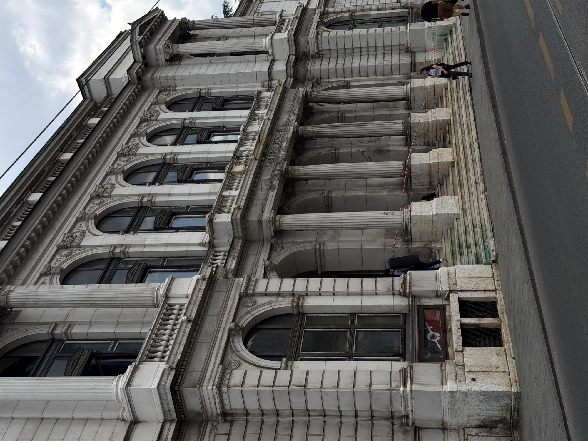 The image shows the National Theatre of Sarajevo, located in Sarajevo, Bosnia and Herzegovina. The building is a grand, neoclassical structure with a prominent facade featuring tall columns, arched windows, and ornate detailing. In front of the building, a sidewalk runs alongside a road. Several people are visible: a person carrying a large, dark case walks up a set of stairs towards the building, another person is walking on the sidewalk, and two individuals are walking away from the building on the sidewalk. Red and white caution tape is visible, indicating a potential construction or cordoned-off area. The sky is overcast with clouds.