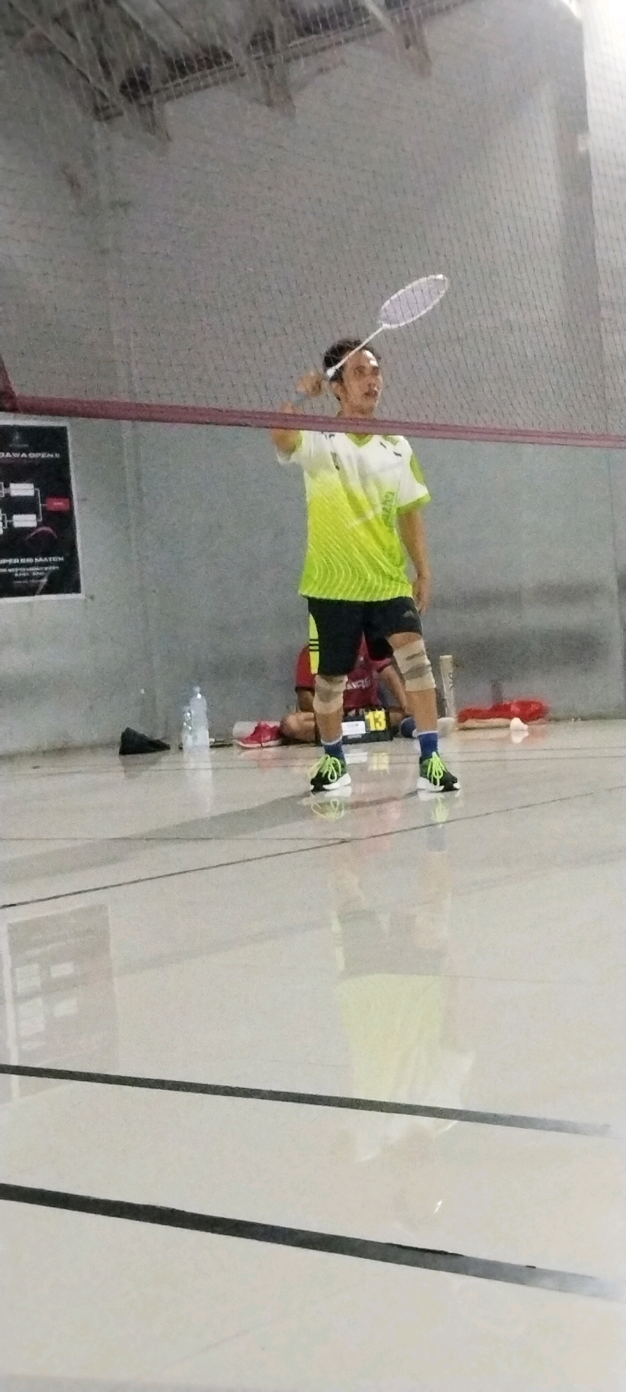 A man, wearing a green and white t-shirt, black shorts, knee pads, and neon green athletic shoes, is captured mid-action playing badminton. He holds a white badminton racket above his head, preparing to hit the shuttlecock. The scene takes place indoors on a polished court with visible court lines. In the background, a net divides the court, and a poster is visible on the wall. A water bottle and other sports equipment are scattered near the wall. The lighting suggests an indoor sports facility. Based on the provided context, the location is Kliris, Indonesia.