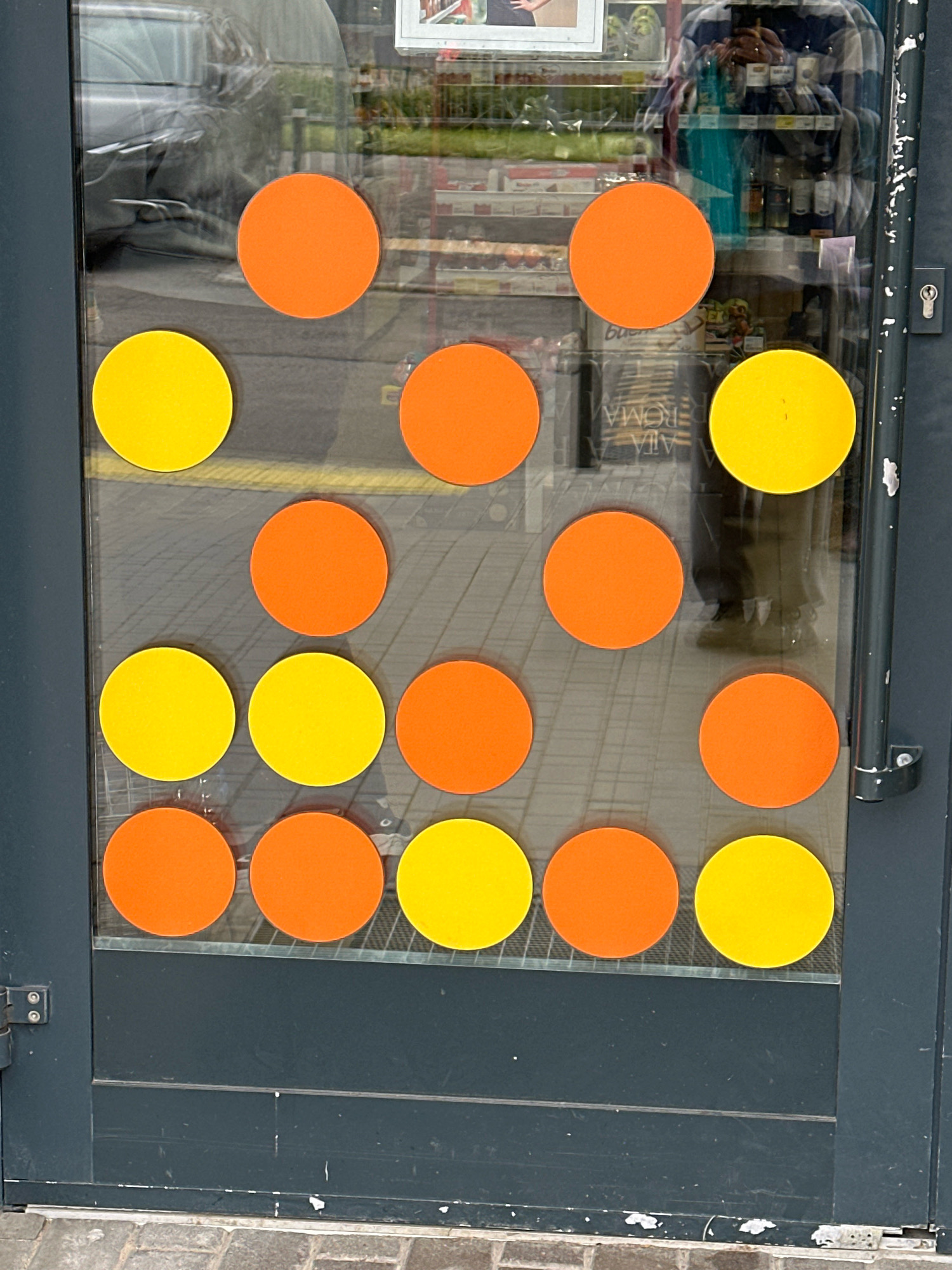 The image shows a glass door with an arrangement of orange and yellow circular stickers. The stickers are arranged in columns and rows, with an alternating pattern of orange and yellow in some columns. Reflections of a person wearing a checkered shirt and what appears to be a store interior are visible on the glass. The door is dark gray with a handle on the right side and hinges on the left. The background suggests an outdoor urban environment with a paved surface and a hint of greenery. The scene is likely captured in the city of Vidnoye, Russia.