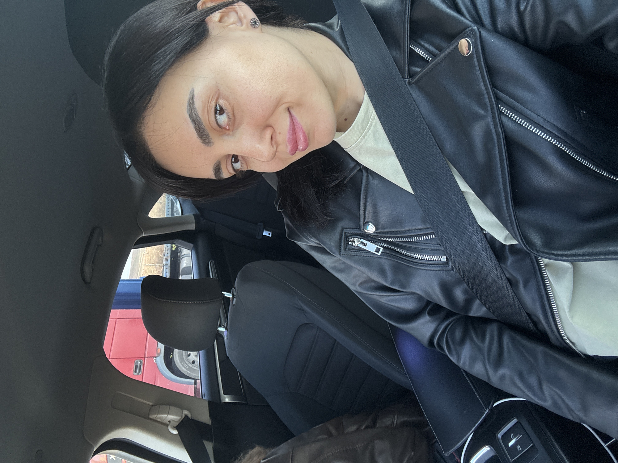A woman with dark hair and fair skin sits in the driver's seat of a car, wearing a black leather jacket over a light-colored shirt and a seatbelt. She's looking at the camera with a slight smile and her tongue visible. Through the car window, a red and blue vehicle is visible, suggesting a street scene. Based on the available information, the location cannot be definitively identified as Tbilisi, Georgia.