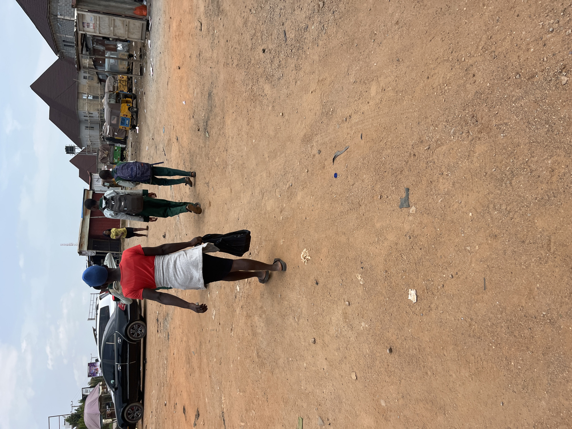 The media file depicts an outdoor scene in Abuja, Nigeria, on what appears to be a clear, overcast day. The setting is a dusty, unpaved area with buildings and vehicles visible in the background.
Several people are present in the image, walking on the dusty ground. In the foreground, a person wearing a red shirt, a white wrap around their waist, and a blue cap is walking away from the camera. They are carrying a black bag in their right hand and appear to be wearing flip-flops. Further in the background, two individuals, likely young males, are walking towards the foreground. One is wearing a light-colored shirt and dark pants with a dark backpack, while the other wears a green shirt and dark pants with a blue and white backpack. In the far background, more people are visible near some stalls or shops, and a young woman in a yellow top stands near a building.
Several objects are scattered throughout the scene, including various vehicles. A black sedan is parked on the left side of the image. Behind it, a yellow auto-rickshaw and a green auto-rickshaw are visible, suggesting a mode of transport common in the region. The buildings in the background appear to be a mix of shops and residential structures, some with corrugated iron roofing and others with more modern construction.
The ground is predominantly sandy and dry, with scattered debris and small rocks. There is no visible text in the image. The overall impression is a snapshot of daily life in a bustling area, likely a market or a busy street in Abuja.