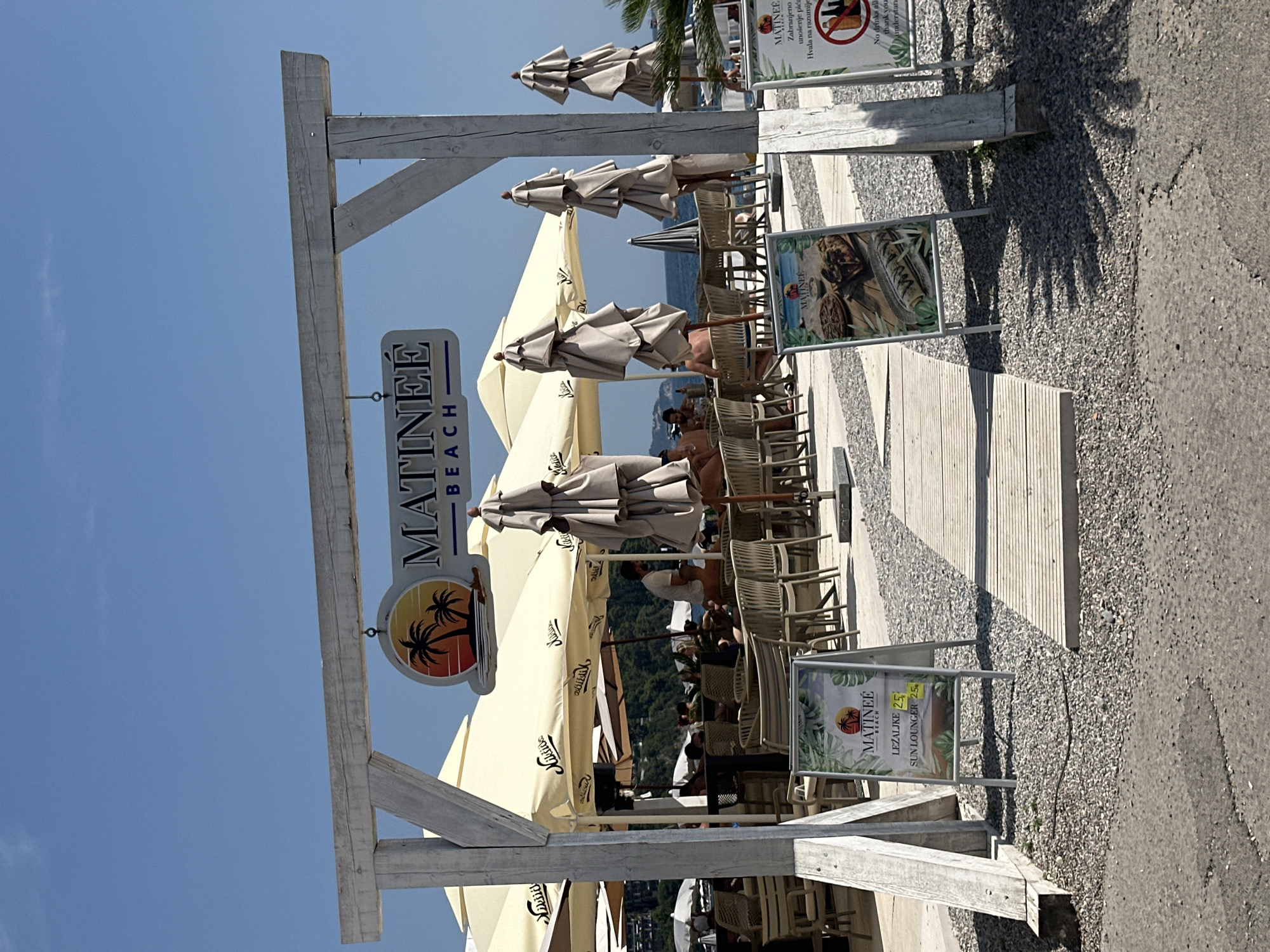 This is a photograph of Matinee Beach in Budva, Montenegro, on a sunny day. The scene depicts an outdoor seating area for a beach establishment. In the foreground, a weathered wooden archway frames the view of the establishment. A sign hanging from the archway reads "MATINEE BEACH" with a graphic of palm trees against a sunset.
Underneath the archway, several beige umbrellas provide shade over tables and chairs where patrons are seated. Some of the umbrellas have the establishment's name, "Matinee," printed on them. People are visible sitting at tables, some appearing to be conversing or relaxing.
To the left and right of the archway, there are A-frame signs. The one on the left displays images of food and text that appears to be a menu or pricing list, including "LEZALJKE" (sun loungers) and "SUN LOUNGER" with prices. The sign on the right has text in a foreign language, with a prominent prohibition symbol against drinking alcohol.
The ground is covered in small, grey pebbles, and a wooden pathway leads further into the establishment. In the background, the sea is visible under a clear blue sky, with distant green hills. The overall impression is of a relaxed beachside cafe or restaurant.