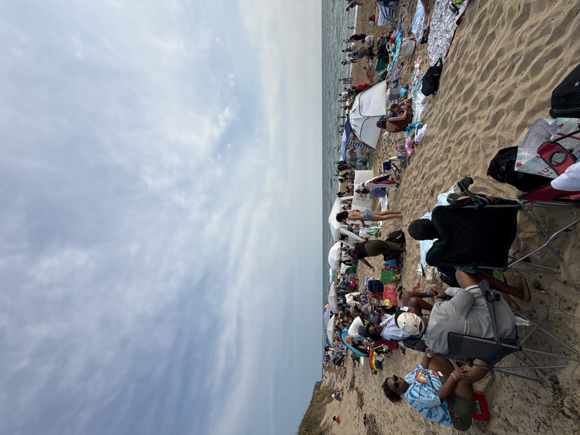 The image depicts a crowded beach scene in Provincetown, Massachusetts, United States of America. The beach is covered in sand and strewn with numerous beachgoers, tents, umbrellas, and beach chairs. The ocean is visible in the background, with waves gently rolling onto the shore. The sky is partly cloudy, with patches of blue sky interspersed with white clouds. People are engaged in various beach activities, such as sitting in chairs, lounging on blankets, and walking along the shore. Some individuals are under the shade of umbrellas and tents. The overall atmosphere appears to be one of leisure and recreation on a summer day.