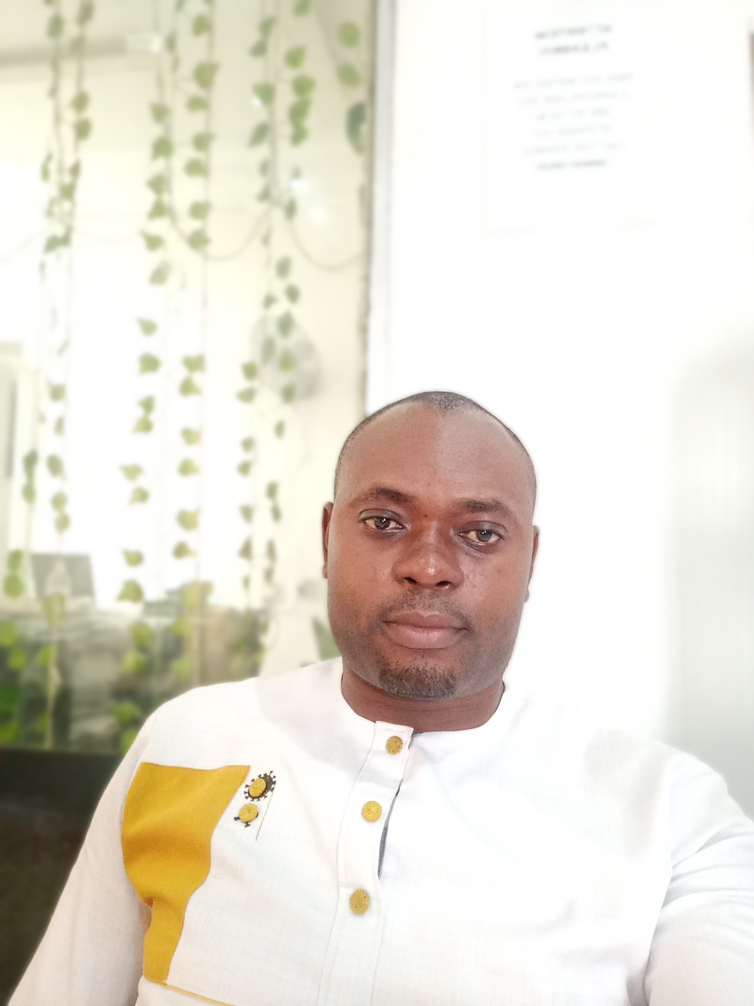 A portrait-style image captures a man of dark complexion looking directly at the camera with a neutral, composed expression. He has short, dark hair and a neatly trimmed beard. He wears a white long-sleeved shirt with a stand collar, adorned with gold-colored buttons down the placket and on the cuffs. A distinct yellow fabric accent is sewn onto the upper left side of his chest, featuring small, dark, circular embroidered patterns.
The background is softly blurred, indicating a shallow depth of field. To the left, artificial green vines with small leaves hang vertically, creating a decorative element. To the right, a white wall or board displays faint, dark, and mostly illegible text; the word "IMPORTANT" might be discernible near the top. The bright, even lighting suggests an indoor daytime setting. The scene is located in Gwarinpa, Nigeria.