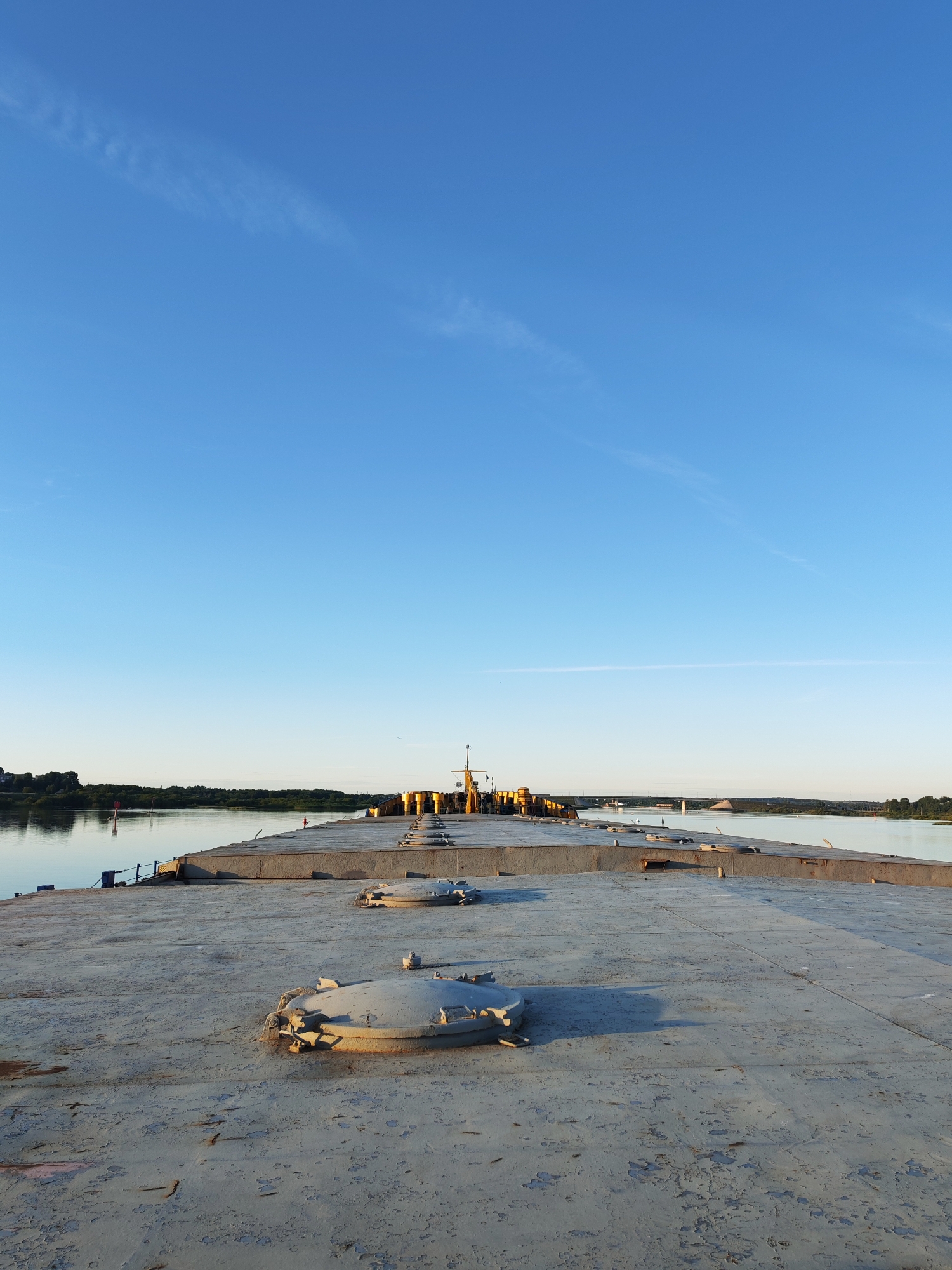 A view from the deck of a barge on a calm river under a clear blue sky. The barge's deck is concrete, weathered and worn, with several circular, metal hatches visible. In the distance, the barge stretches out, lined with yellow cylindrical objects, possibly part of its cargo or equipment, and a yellow crane. The river, reflecting the sky, is bordered by a lush green tree line on the far bank. A few small structures and a red buoy are visible on the distant shore. The sun is low in the sky, casting a warm light that illuminates the scene, suggesting it's either early morning or late evening. The overall mood is peaceful and still. The location context is Нифантово, Russia.