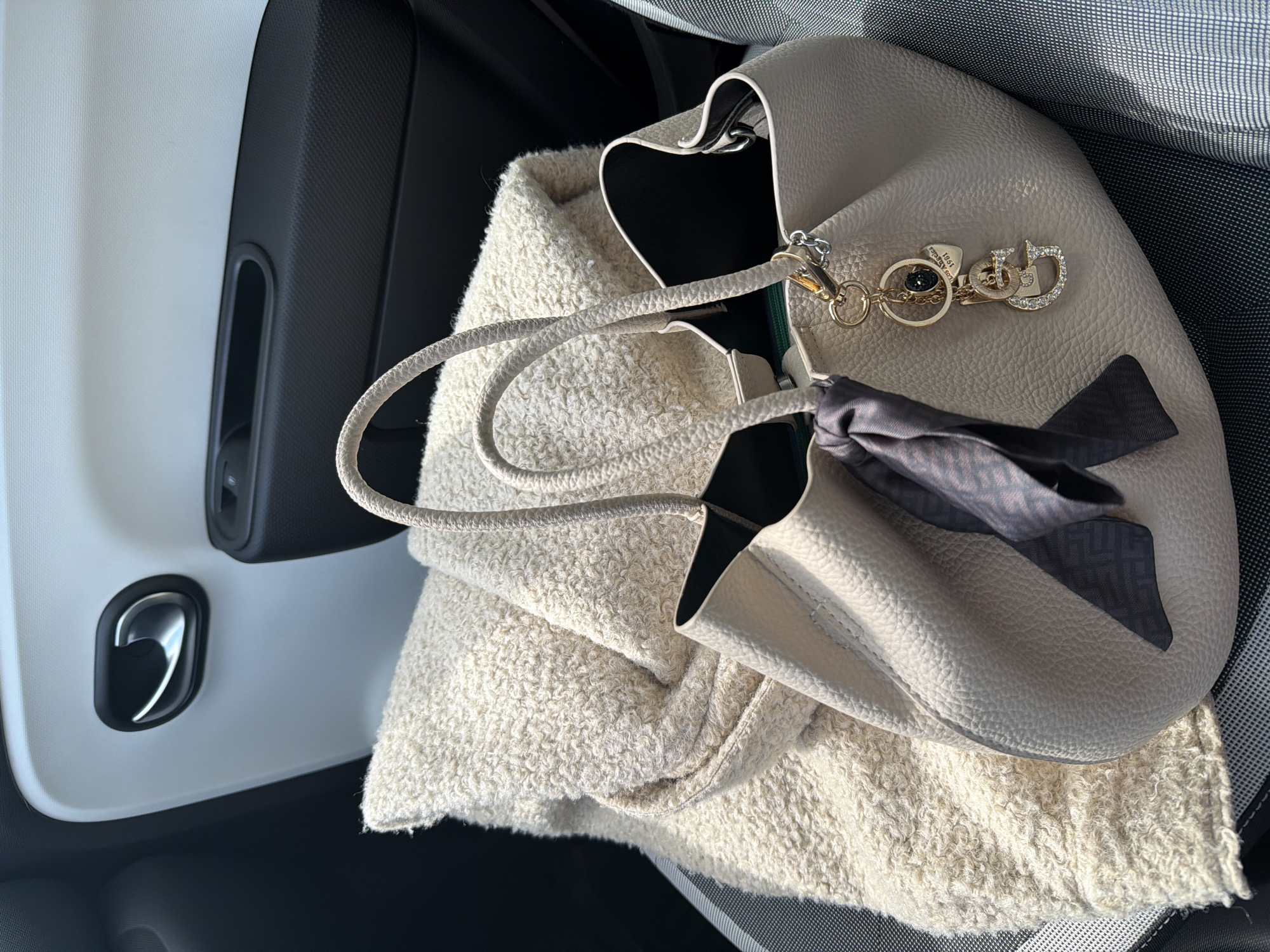 The image displays the interior of a vehicle, specifically focusing on the passenger seat and an adjacent door panel, consistent with a Renault Twingo III. A light beige, textured handbag rests on a grey woven-patterned car seat. The handbag features two top handles, a dark grey silk twilly with a repeating logo pattern tied around one handle, and a gold-toned keychain. The keychain components include a large, gemstone-encrusted "G" initial, a smaller "G" initial, a heart-shaped charm inscribed "Los Angeles 1981," and a small black bead charm. Adjacent to the handbag, a folded, cream-colored, bouclé-textured blanket or garment is visible. The car door panel in the background is two-toned, featuring a white upper section and a black textured lower section, with a chrome interior door handle and a black power window switch present. This scene is located in Sofia, Bulgaria.