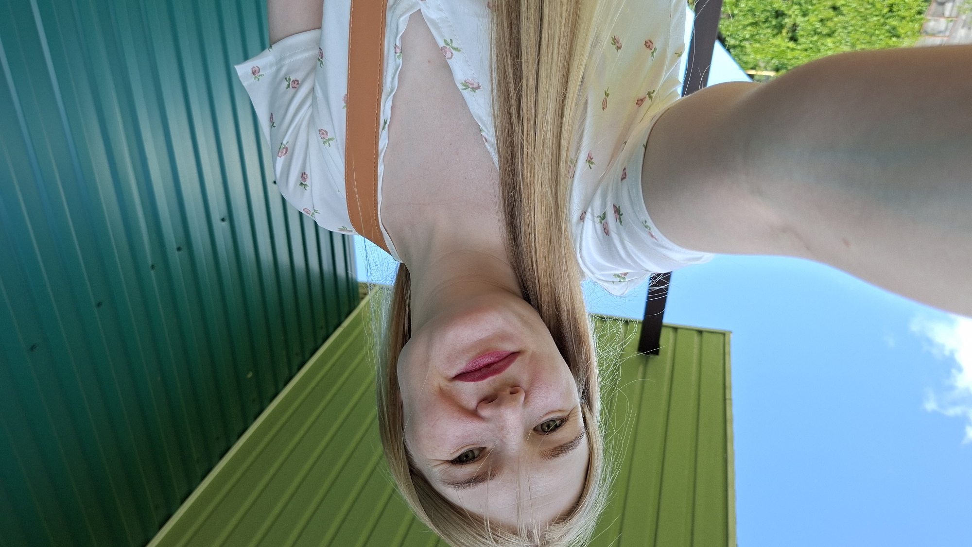 A woman with blonde hair and fair skin is taking a selfie outdoors. She is wearing a white shirt with a floral pattern and has on a brown strap across her chest. She is smiling and looking at the camera. The background consists of a blue sky with some clouds, a green corrugated metal wall, and a green overhang. The image appears to be taken in a rural or suburban setting, possibly in Большой Букор, Russia, given the presence of a typical dacha-style overhang and building. There is greenery visible in the lower left corner, suggesting vegetation.