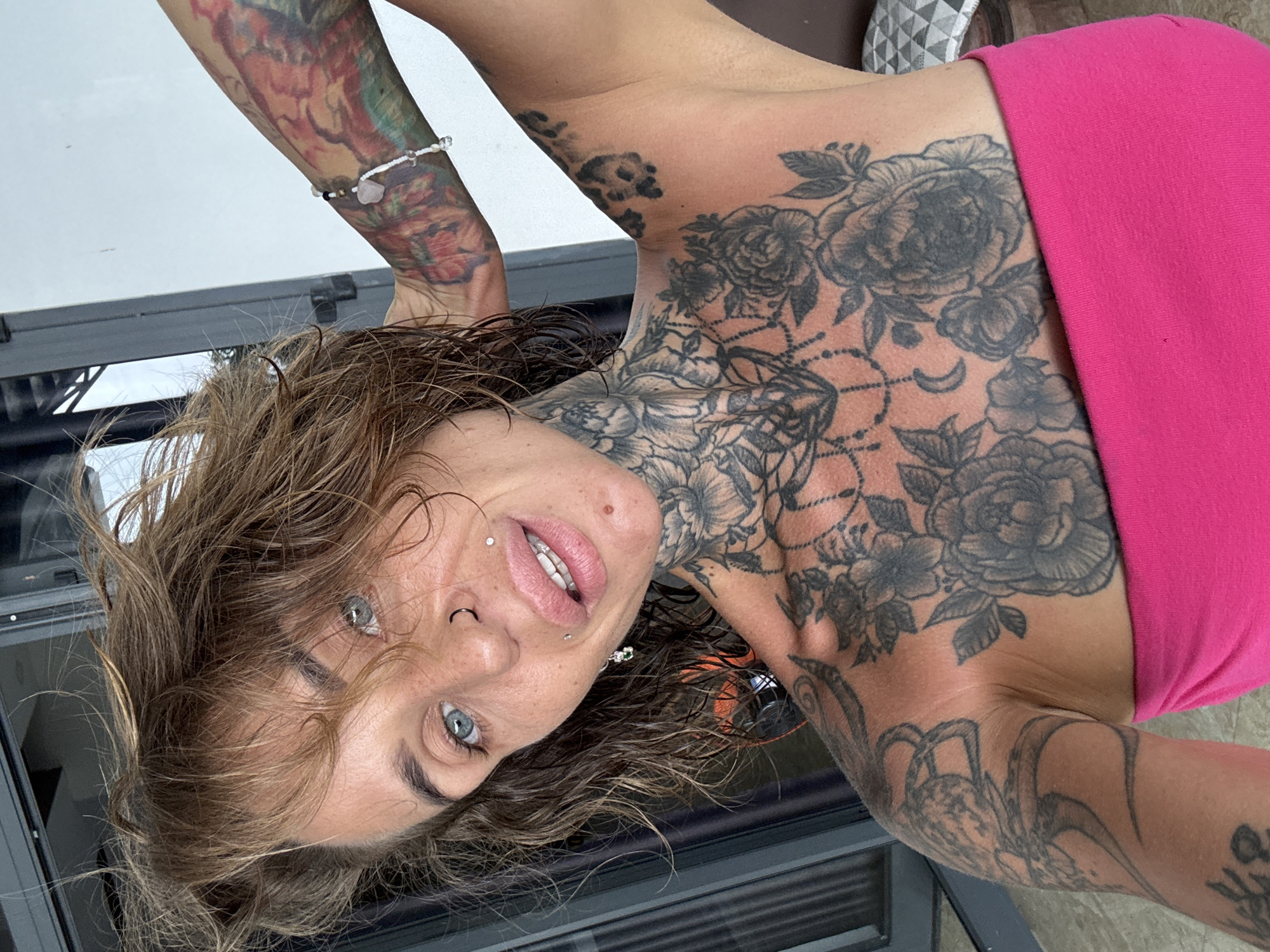 A woman with wet, tousled brown hair and blue eyes is looking at the camera. She has several facial piercings, including a septum ring and dermal piercings on her lips. Her neck and chest are covered in intricate black tattoos, and her upper arm also has tattoos. She is wearing a bright pink strapless top. The background appears to be outdoors, with a hint of a building with dark window frames and some reflective surfaces. The lighting suggests it might be daytime. The image is a close-up portrait, focusing on the woman's face and upper body. The provided information about the location being Bac Nha Trang Ward, Vietnam is not visually verifiable from the image itself.
