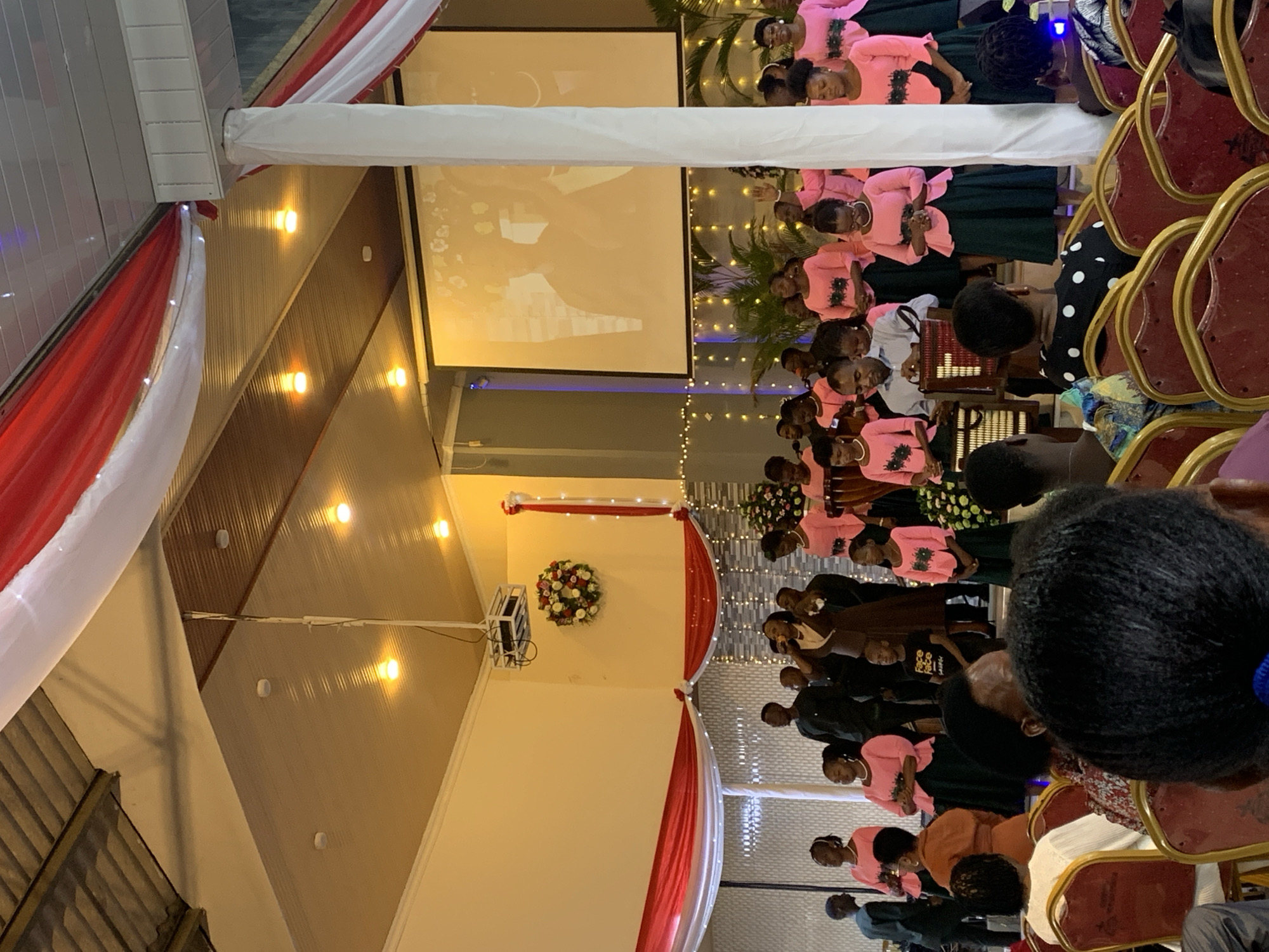The image depicts an indoor gathering in a room decorated with red and white draping, possibly for an event or ceremony. A projector screen displays an image, and there are rows of people seated on red chairs facing the screen. Many individuals are wearing pink tops with dark green skirts. Based on the location of the prompt, this event is likely taking place in Dar es-Salaam, Tanzania.