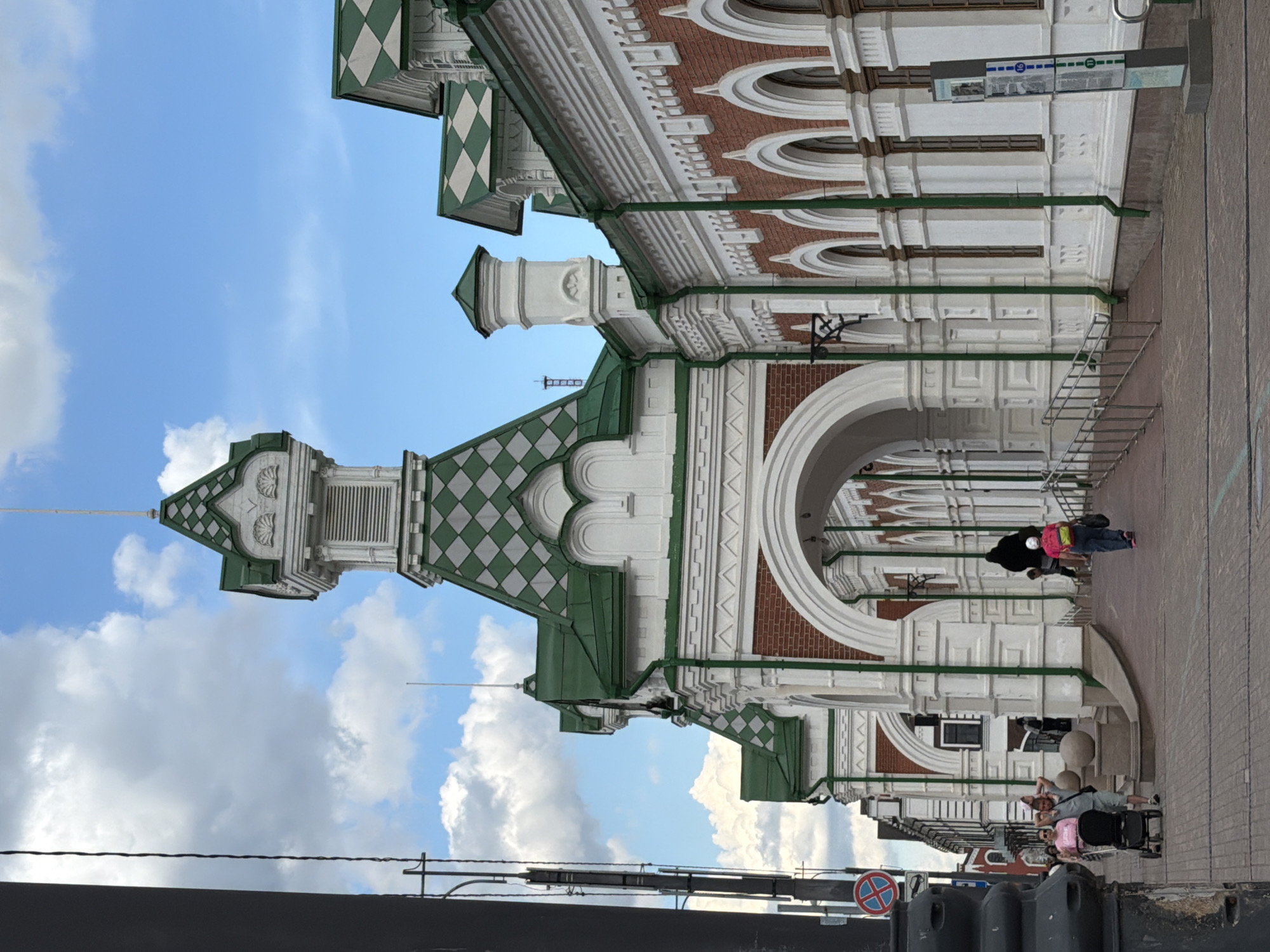 The image depicts a street scene in the city of Perm, Russia, featuring a historic building with a distinctive green and white diamond-patterned roof and ornate white architectural elements. The building has a brick facade with arched windows. In the foreground, a paved walkway is visible, with several people walking and one person pushing a stroller. The sky above is partly cloudy. The overall impression is of a pleasant day in an urban environment with notable architecture.