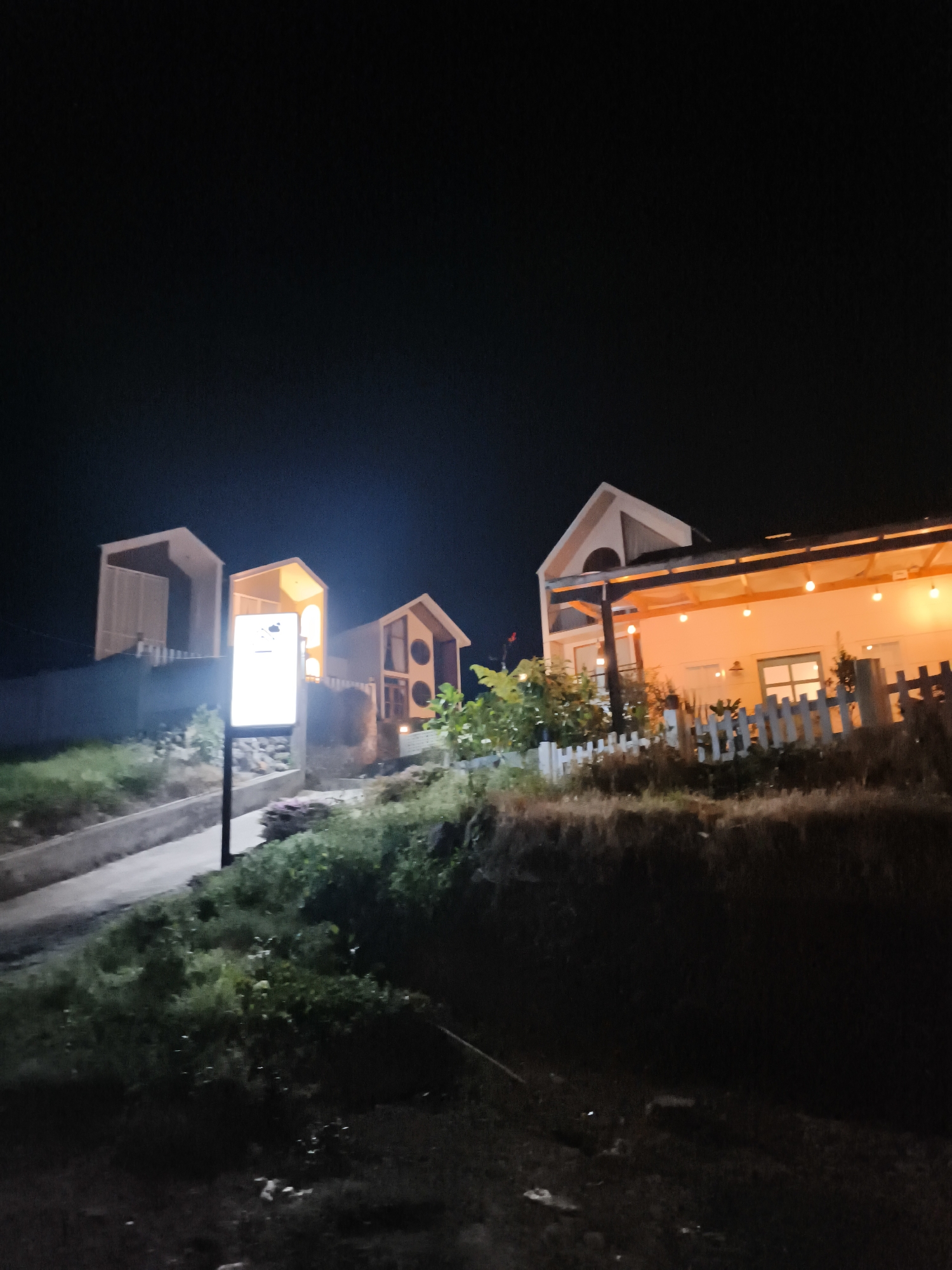 The image shows a nighttime exterior scene featuring several small, modern structures with peaked roofs. The structures are illuminated, with some featuring warm, orange lights. One structure has a covered porch area with multiple bulbs. A white picket fence is visible in front of some of the buildings. In the foreground, there is a pathway or road leading uphill, bordered by green vegetation. A lit signpost stands to the left of the pathway. The overall impression is of a small cluster of buildings, possibly accommodations or a business, set against a dark, natural environment. The location is identified as Dieng Kulon, Indonesia.