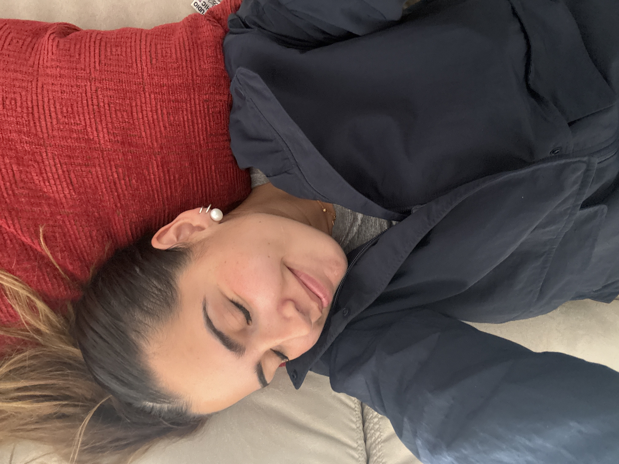 An indoor, eye-level shot captures a woman lying on her side, resting her head on a textured red pillow. She is positioned horizontally in the frame, with her head towards the left. Her eyes are closed, and a gentle smile plays on her lips, conveying a sense of peace or contentment.
The woman has dark hair that transitions to a lighter, caramel blonde towards the ends. She is wearing a dark blue jacket or coat over what appears to be a grey top. A large white pearl earring, surrounded by smaller silver rings or hoops, is visible on her left ear. A delicate gold chain necklace with a small pendant can be seen around her neck, peeking out from under her jacket. Her skin appears natural with a slight flush.
She is resting on a beige or light brown upholstered surface, likely a sofa or bed. The red pillow, which has a distinct corduroy-like texture, supports her head. On the pillow, a small white fabric tag is partially visible with the text "STUDIO RIC". The lighting suggests an indoor setting, and no specific time of day or weather conditions are discernible from the image. The scene conveys a moment of relaxation. The location context provided is Santa Clara Ocoyucan, Mexico.