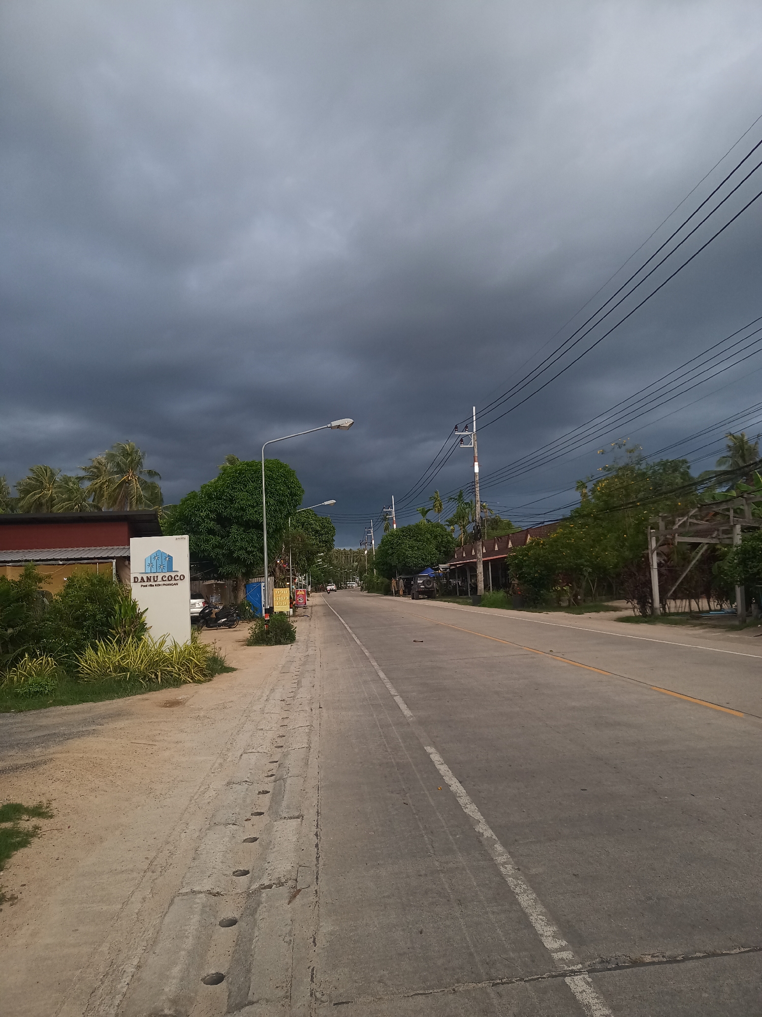 The image depicts a street scene in Moo 4, Thailand, characterized by overcast skies and a sense of impending rain. The road, made of gray asphalt with a double yellow center line and a broken white line marking the edge of the lane, stretches into the distance. On the left side of the road, a white sign with the words "DANU COCO Pool Villa KOH PHANGAN" is visible. Vegetation is abundant on both sides of the road, with trees and bushes lining the sidewalks and yards. Power lines crisscross above the street, supported by utility poles. The environment suggests a rural or semi-rural area. The image is devoid of people, but there are motorcycles parked behind the sign.