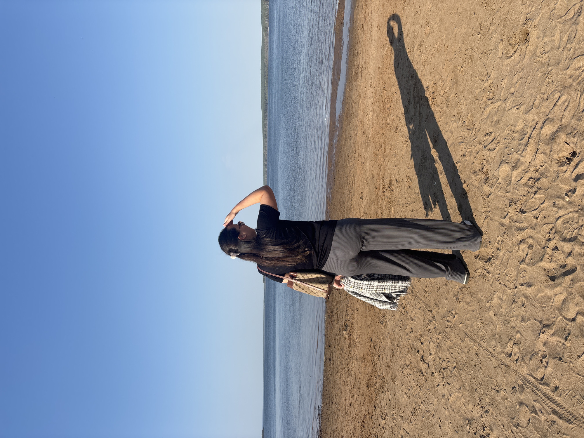 The image shows a person standing on a sandy beach near water. The person is wearing a black shirt and striped pants. Their right arm is raised and their hand is touching their head. A handbag is slung over their left shoulder and there is a patterned blanket. The shadow of the person and their raised arm can be seen on the sand.
The location is Chaikovsky, Russia.