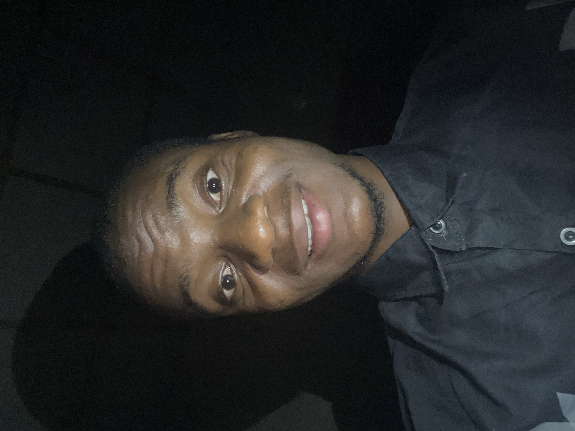 A medium close-up photograph shows a smiling dark-skinned man. He is wearing a black collared shirt with button fasteners. The background is dark and indistinct. It is not possible to verify that the photograph was taken in Minna, Nigeria based on the available information.