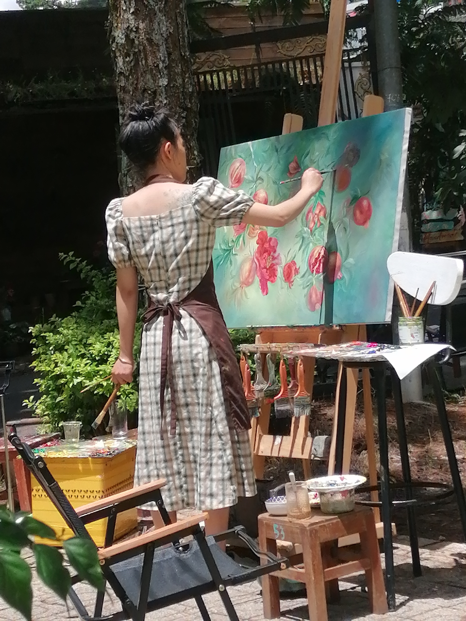 An artist, a woman with her hair in a bun, wearing a plaid dress and a brown apron, stands outdoors in front of an easel painting a canvas depicting red fruits and flowers against a teal background. She holds a paintbrush in her right hand, actively applying paint to the canvas. In front of the easel, a wooden table displays an assortment of paintbrushes and a palette filled with various colors. To the left, a yellow chest with a patterned design sits near a folded black chair. The background features lush green foliage and a rough-barked tree. This scene is likely situated in Da Lat, Vietnam, a city known for its artistic community and natural beauty, often hosting artists working outdoors.