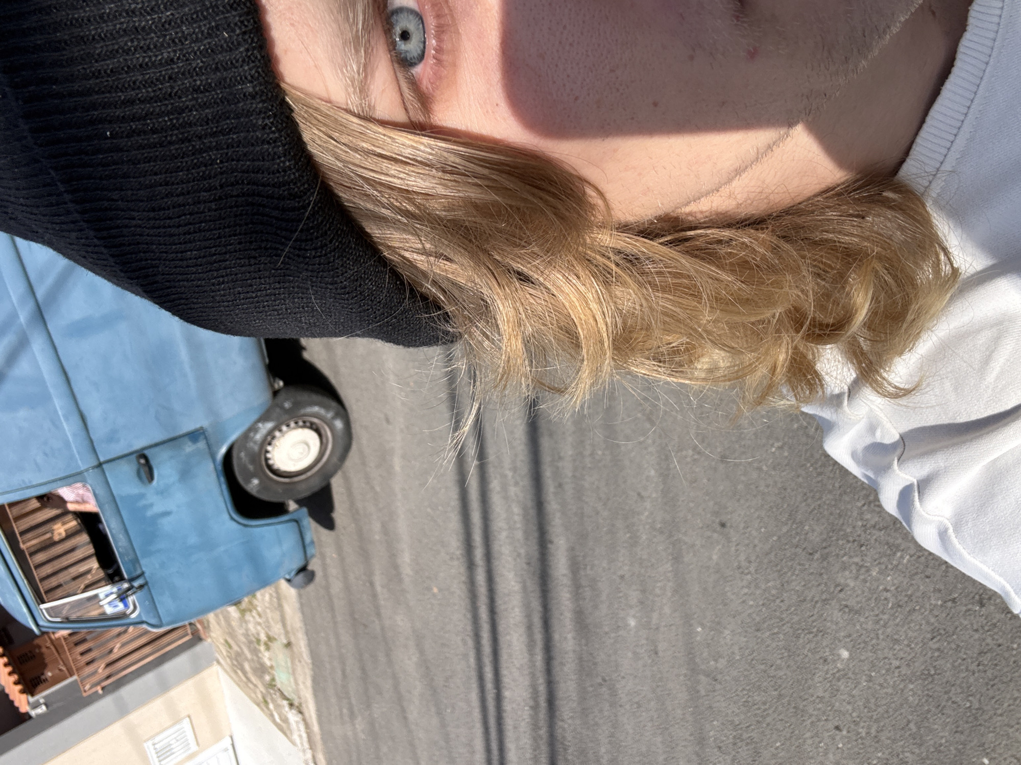 The image is a close-up, outdoor, daytime selfie taken at an angle. A person with blonde hair, wearing a black beanie and a white t-shirt, is partially visible in the foreground. Their blue eye is in focus, and a shadow crosses their face. In the background, a faded blue Volkswagen van is parked on a paved street. The van has a brown fence and a building with a terracotta roof behind it. The setting appears to be a residential street, possibly in São Paulo, Brazil, given the architectural style of the building and the presence of overhead wires casting shadows on the street.
