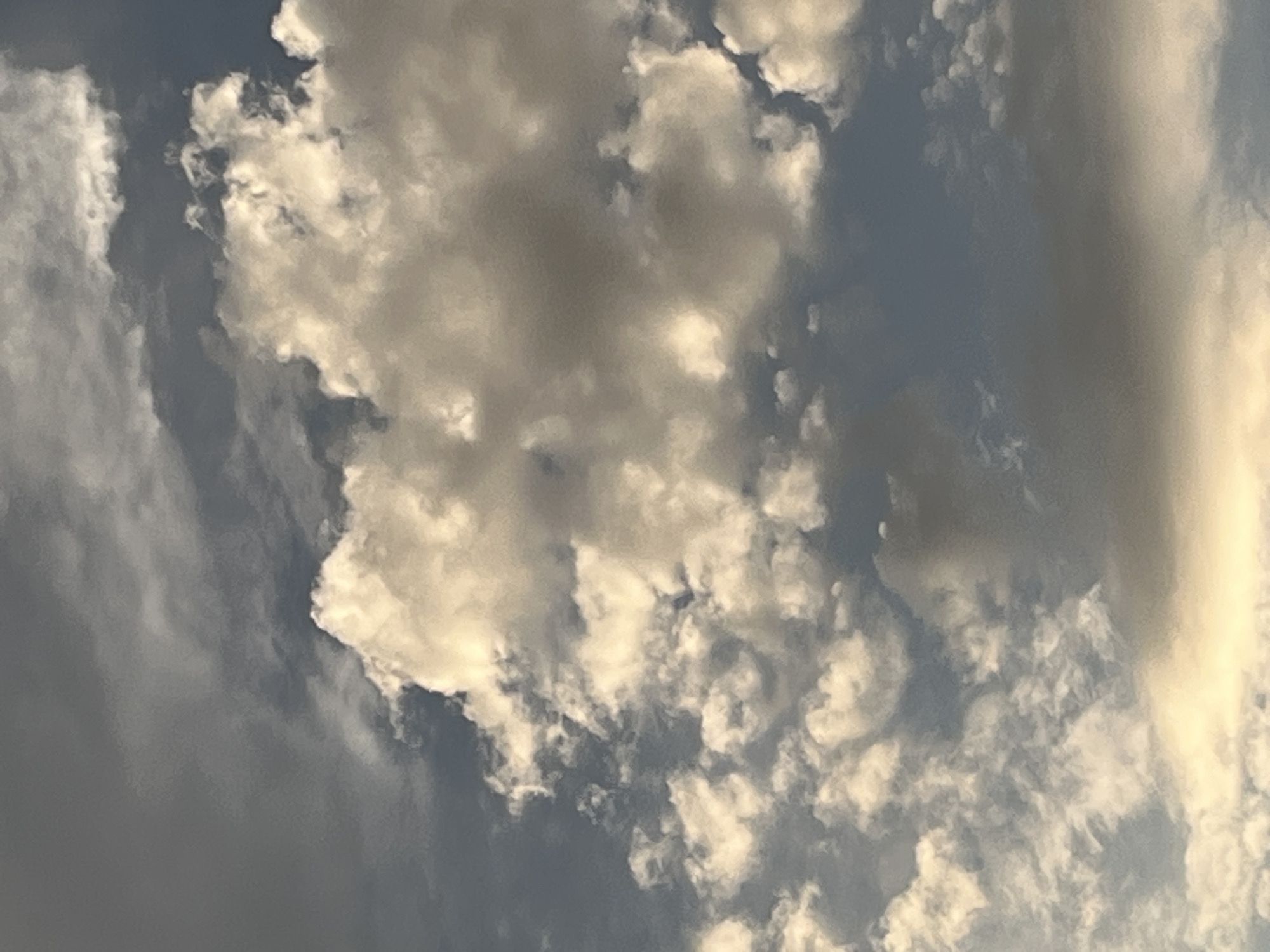 The image captures a sky filled with a mix of dark gray and bright white clouds. The lighting suggests it is either early morning or late afternoon, with sunlight illuminating some of the clouds from behind, creating a dramatic contrast. The blue sky is visible in patches between the clouds. The scene appears to be an outdoor environment, possibly looking upwards from ground level in a city. There are no people, subjects, objects, or specific actions depicted in the image. The location in Krasnyye Barrikady, Russia, is not discernible from the visual content of the photograph.