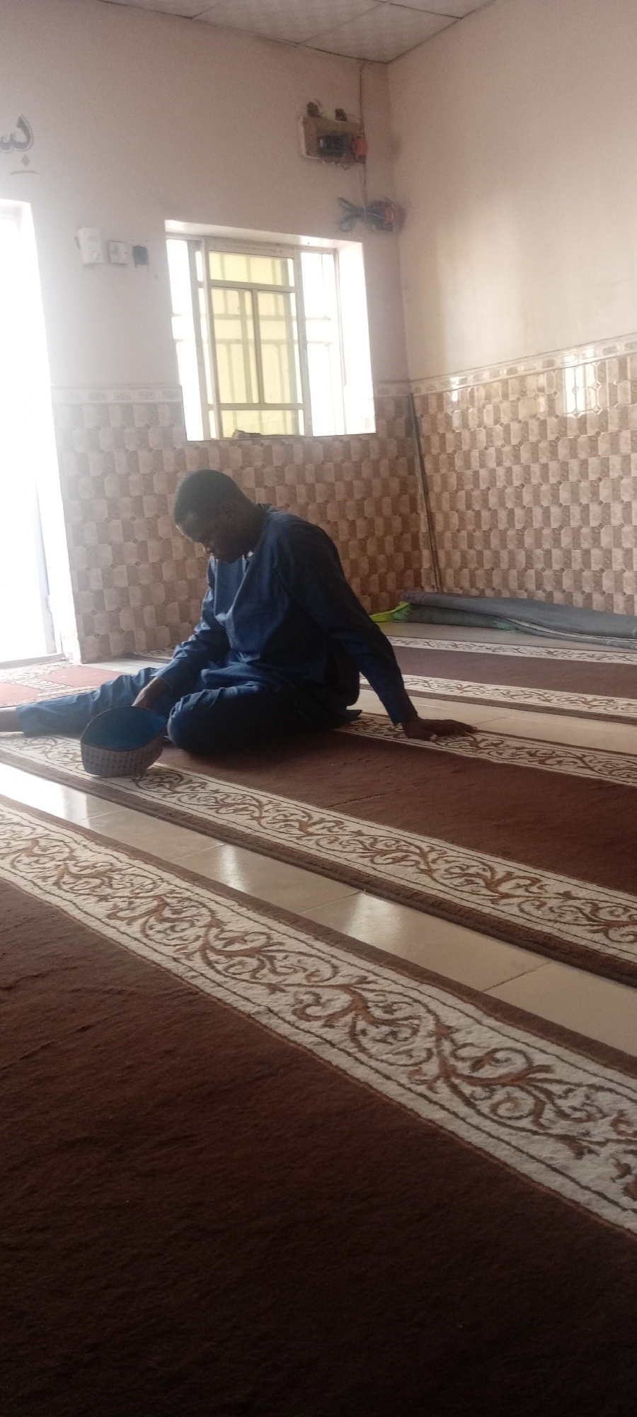 A man in a blue tunic sits on a brown carpet in a room with tiled walls. The carpet is patterned with light-colored, ornate stripes. A light-colored window is visible in the background. He is also wearing a taqiyah. The room appears to be a prayer area. The location may be a mosque in Kano, Nigeria.