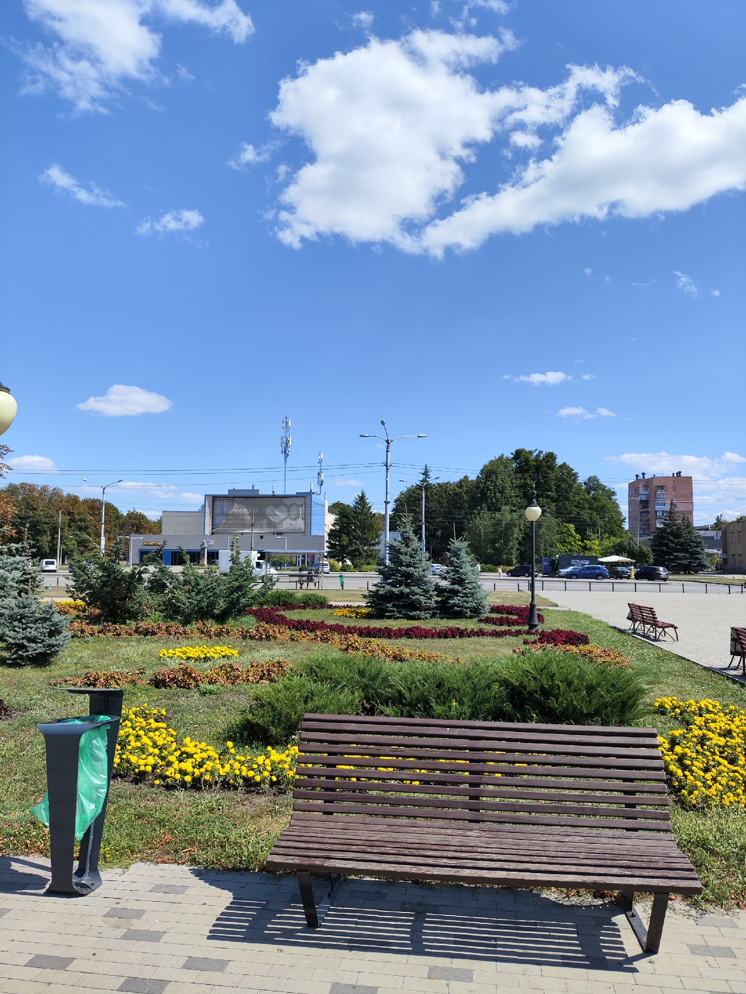 The image captures a public park area on a clear, sunny day. The foreground features a wooden park bench casting a striped shadow on the paved walkway. To the left of the bench is a black trash receptacle with a green trash bag inside. Behind the bench and trash can, a meticulously maintained flower bed displays a vibrant mix of yellow and reddish-brown flowers and green foliage, bordered by manicured bushes and small evergreen trees.
In the mid-ground, a street runs parallel to the park, with several cars parked or driving. Beyond the street, various buildings are visible, including a modern-looking building with a large display screen or billboard, a taller brick building, and other structures partially obscured by trees. Several cellular antennas are mounted atop one of the buildings. The background is filled with lush green trees and a bright blue sky dotted with fluffy white clouds. This scene is located on the corner of Sumska Street and Constitution Square in Kharkiv, Ukraine.