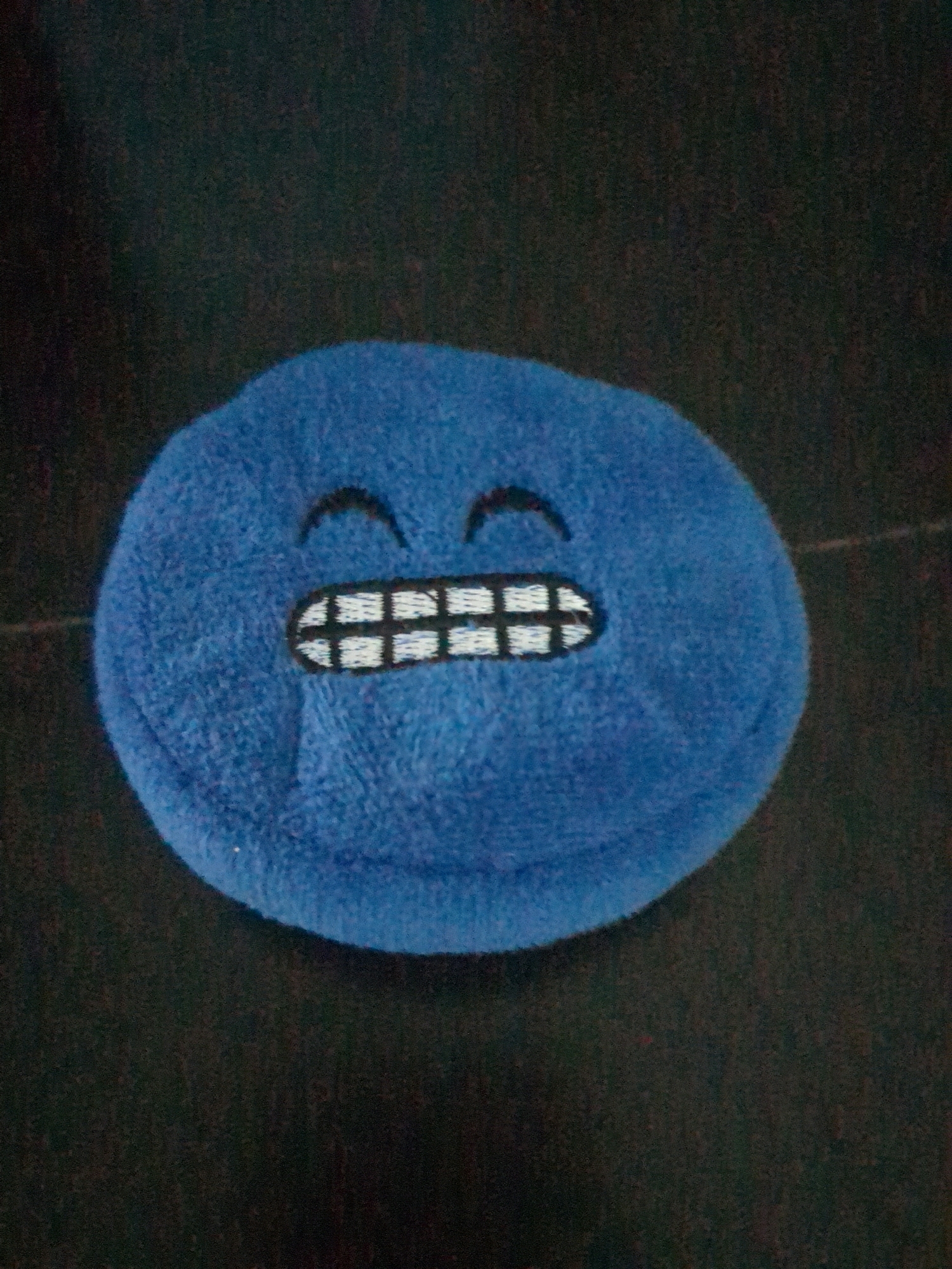 A blue, plush, circular object with embroidered facial features sits on a dark, textured surface. The object has two black, crescent-shaped eyes and a wide, white mouth with horizontal black lines representing teeth. The surface it rests on appears to be fabric, possibly a blanket or clothing. There is no indication of a specific city or location within the image.