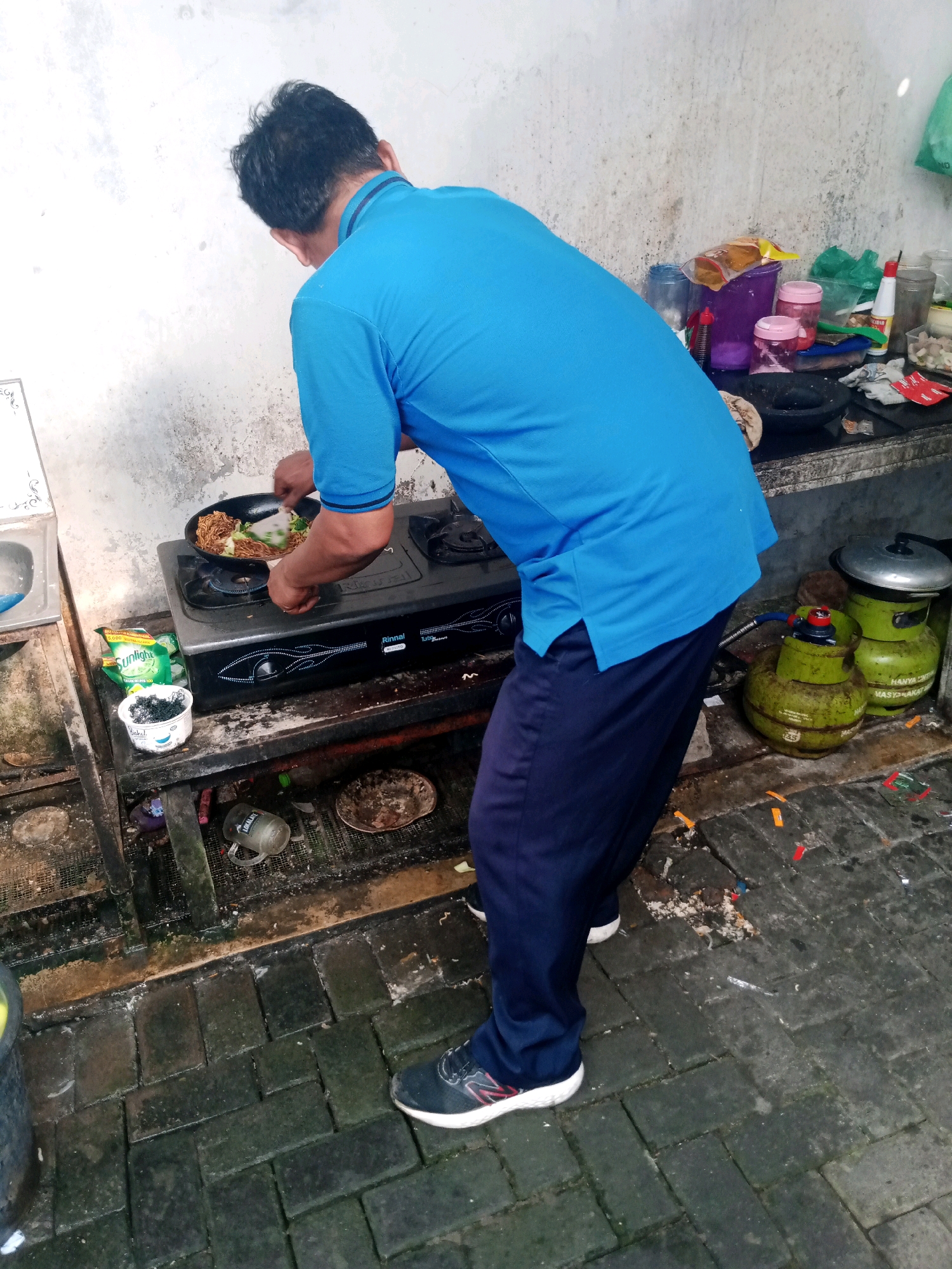 The image depicts an outdoor cooking scene in Surakarta, Indonesia. A man, wearing a blue polo shirt and dark pants, is standing and actively cooking. He is using a black two-burner gas stove, which is placed on a rustic wooden shelf. In a pan on the stove, he is stirring noodles and vegetables with a spatula. To the left of the stove, there is a small white bowl containing some dark, possibly chopped, ingredients, and a green packet of Sunlight dish soap. To the right of the stove, on the same shelf, are various containers, including a purple container, pink jars, and a white bottle with a red cap. Further to the right, two green propane gas cylinders are visible, one connected to the stove. The ground is paved with dark bricks and appears wet, with some scattered debris. The background features a textured, light-colored wall, possibly concrete or plaster, showing signs of age and discoloration. The overall environment suggests a casual, functional outdoor cooking area, likely in a residential or informal commercial setting within Surakarta.