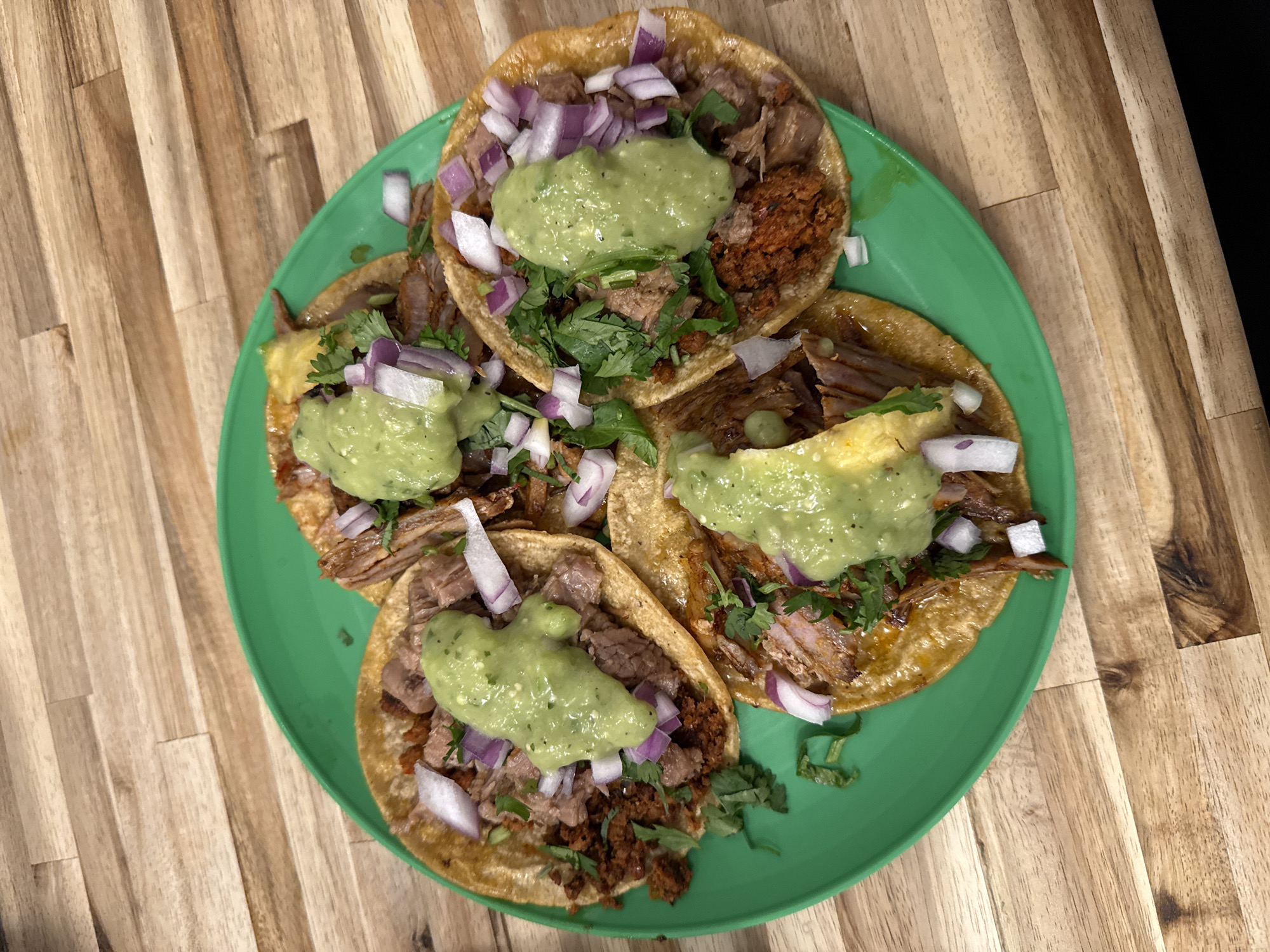 The image shows a green plate of four tacos on a wooden surface. The tacos appear to be filled with meat, a green sauce (possibly guacamole or a similar avocado-based sauce), chopped red onion, and cilantro. The scene seems to be indoors, potentially in a restaurant or kitchen setting. There is no evidence to determine the location in the city of New York, United States of America.